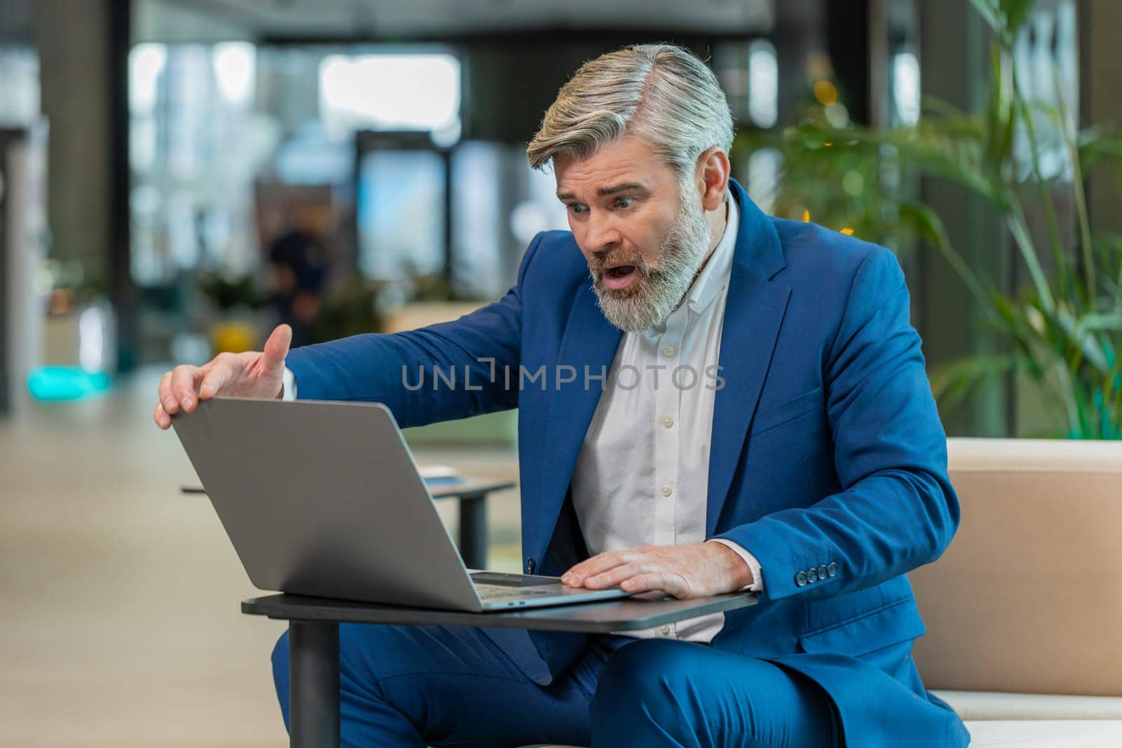 Sad middle-aged Caucasian businessman using laptop typing browsing, loses becoming surprised sudden lottery results, bad news, fortune loss fails office lobby. Upset freelance worker in hotel hall.