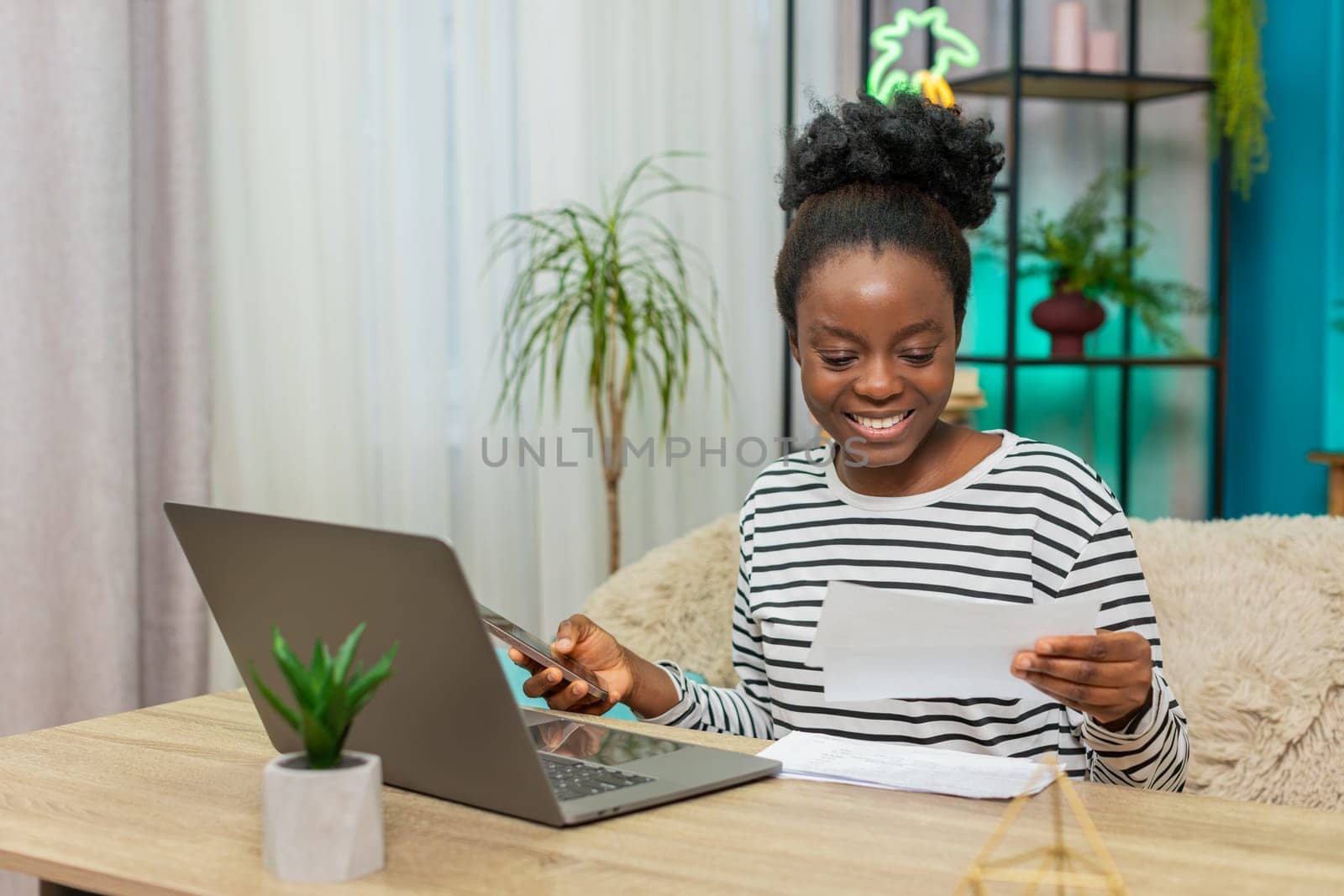 African American woman working on family finances. Girl sitting on home sofa using calculator reviewing electronic bills on laptop. Focused expression occasionally sighing, indoor financial management