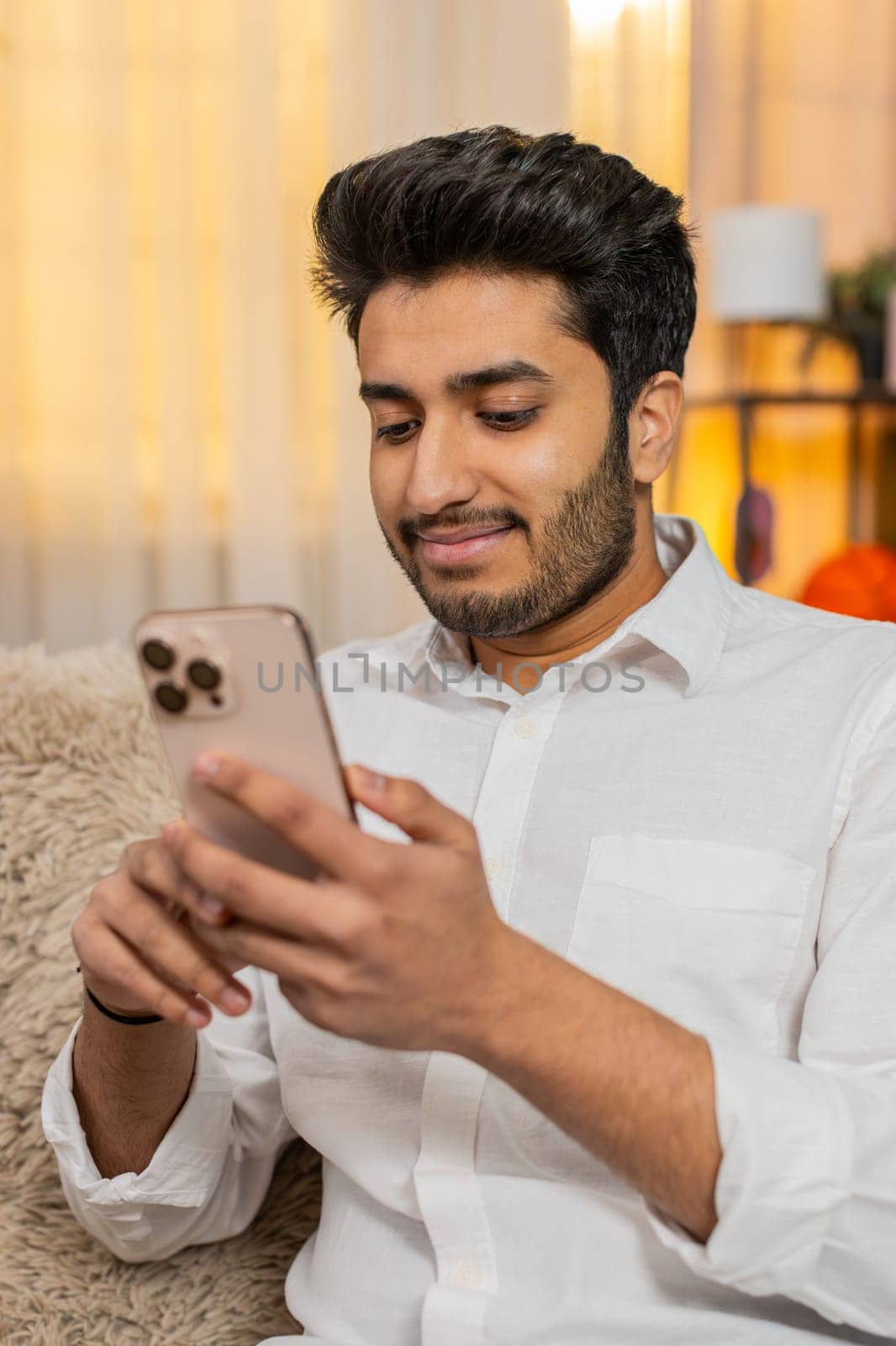 Indian young man at home sitting on sofa focused on smartphone typing text, browsing news, reacting to notifications. Arabian guy using mobile phone for social media, work, texting, digital lifestyle