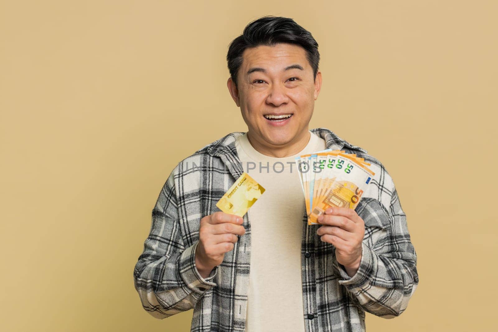 Happy Asian middle-aged man showing plastic credit bank card and money euro cash advertising taking loan financial help for purchases online. Mature Chinese guy isolated on beige background indoors
