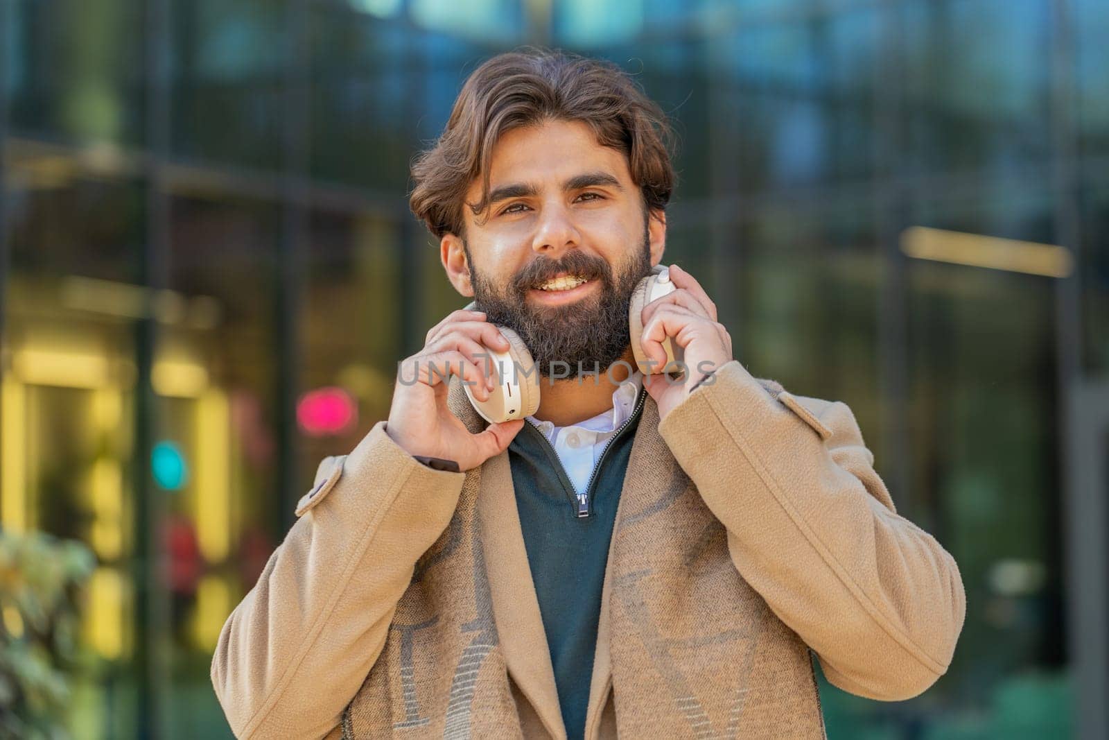 Joyful relaxed overjoyed Arabian business man listening favorite energetic disco rock n roll music outdoors. Adult guy taking off wireless headphones looking at camera smiling on downtown city street.