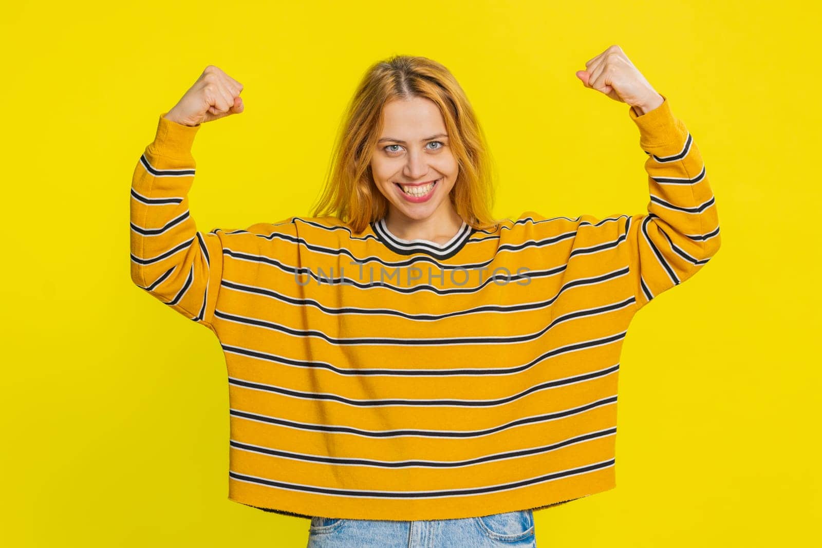 I am strong and independent. Happy smiling Caucasian woman showing biceps and looking confident, feeling power strength to fight for rights, energy to gain success win. Girl on yellow background