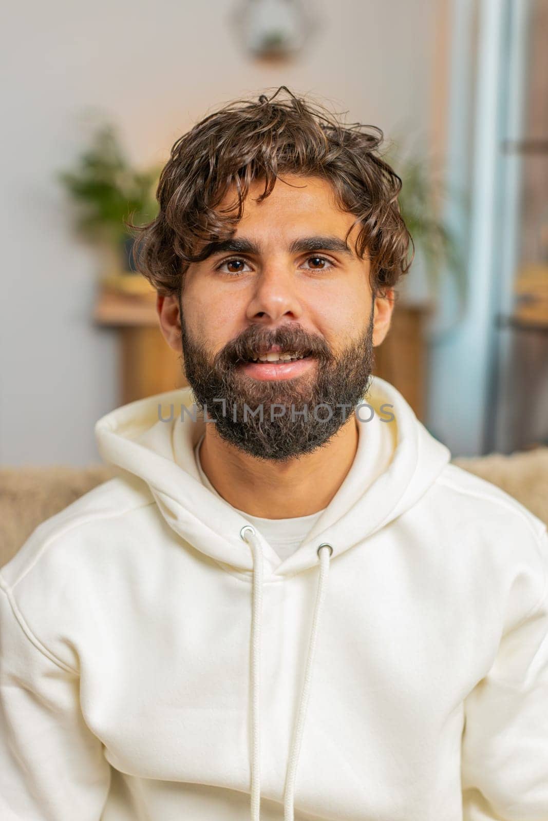 Portrait of smiling young Indian man looking at camera. Happy content adult bearded Hispanic guy in hoodie relaxing feeling satisfied concept sitting on sofa couch in living room at home. Lifestyles.