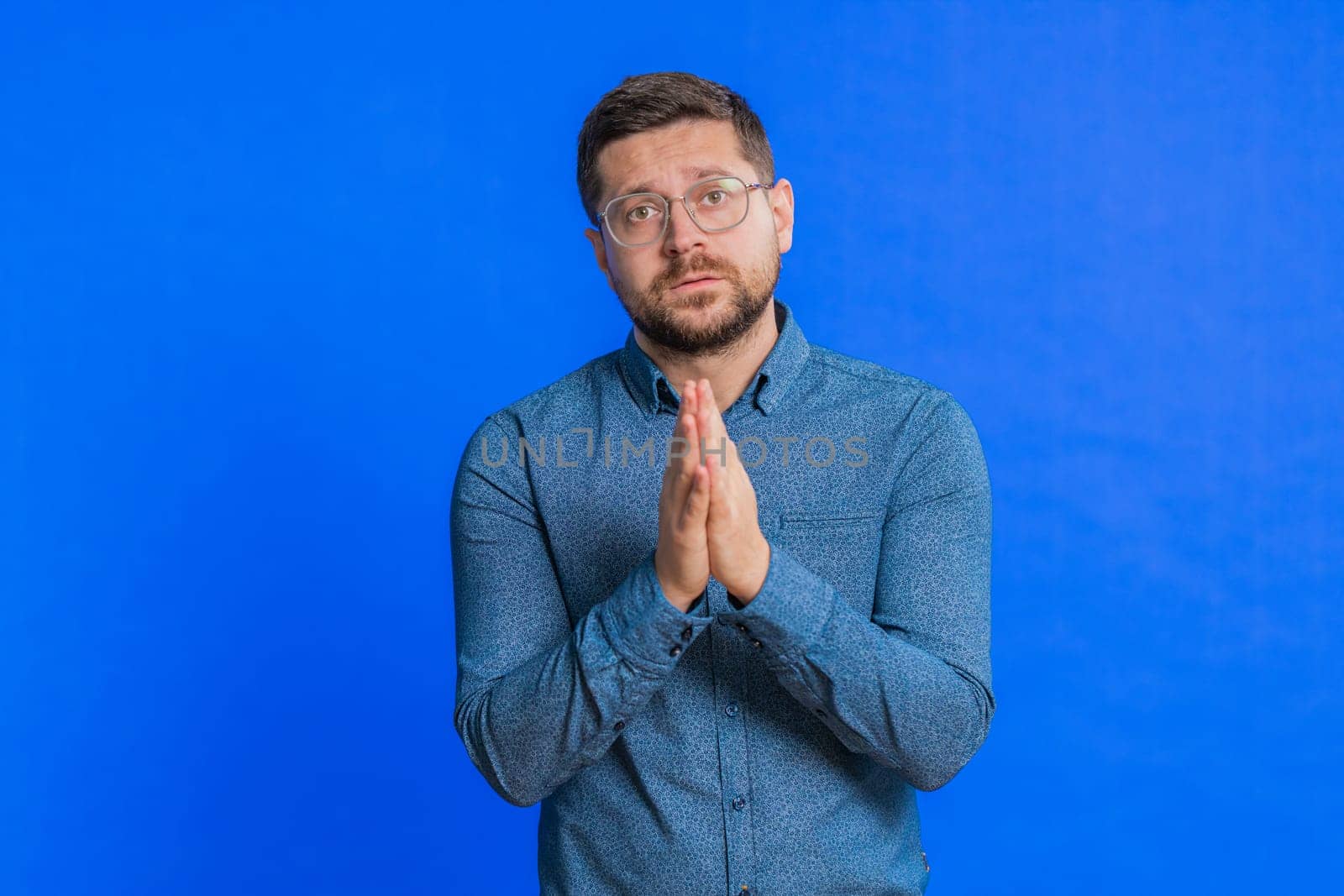 Please, God help. Caucasian middle-aged man praying, looking at camera and making wish, asking with hopeful imploring expression, begging apology, meditation. Adult guy isolated on blue background