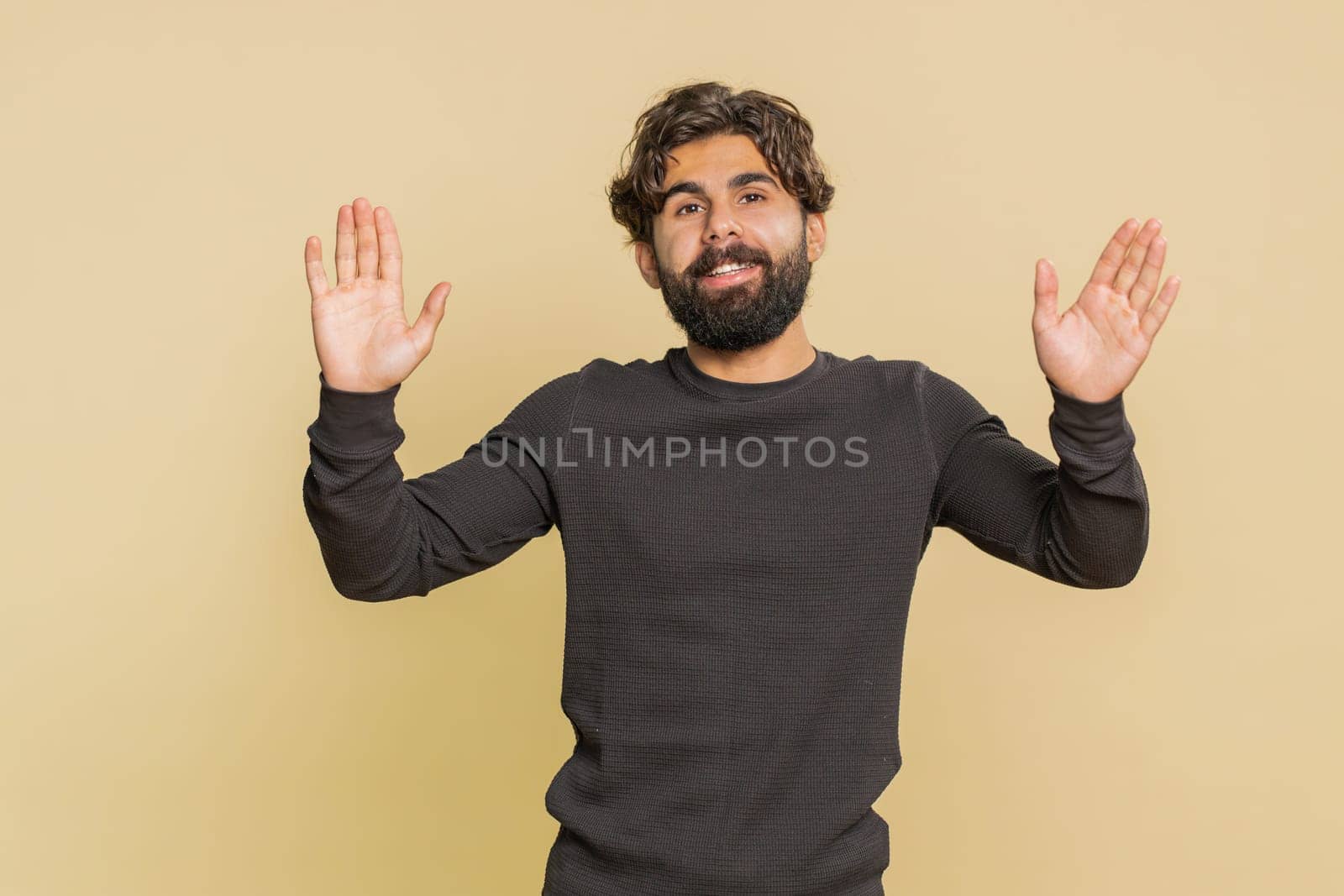 Hello, welcome. Handsome young Indian man with long sleeves waving hi with his palm, greeting with hospitable friendly smile. Adult male Hispanic guy isolated on beige studio background indoors.