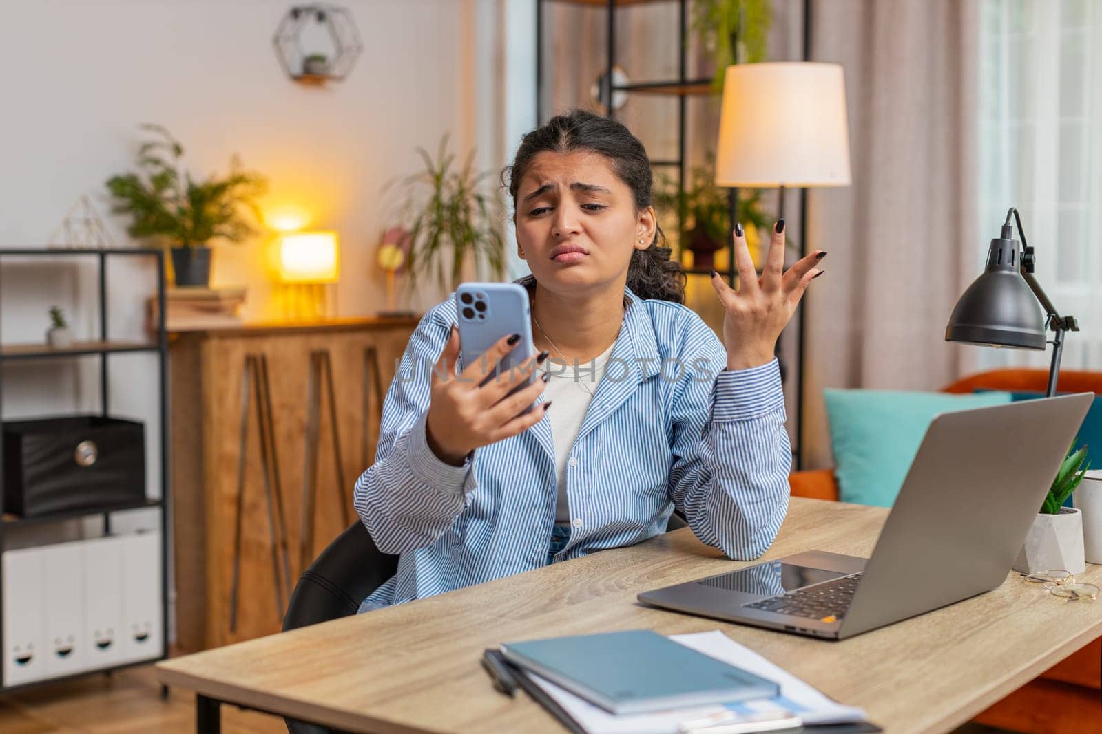 Upset displeased Indian woman freelancer using smartphone sitting at desk table in living room. Sad young girl touching head loses becoming surprised by sudden lottery results or bad news. Lifestyle.