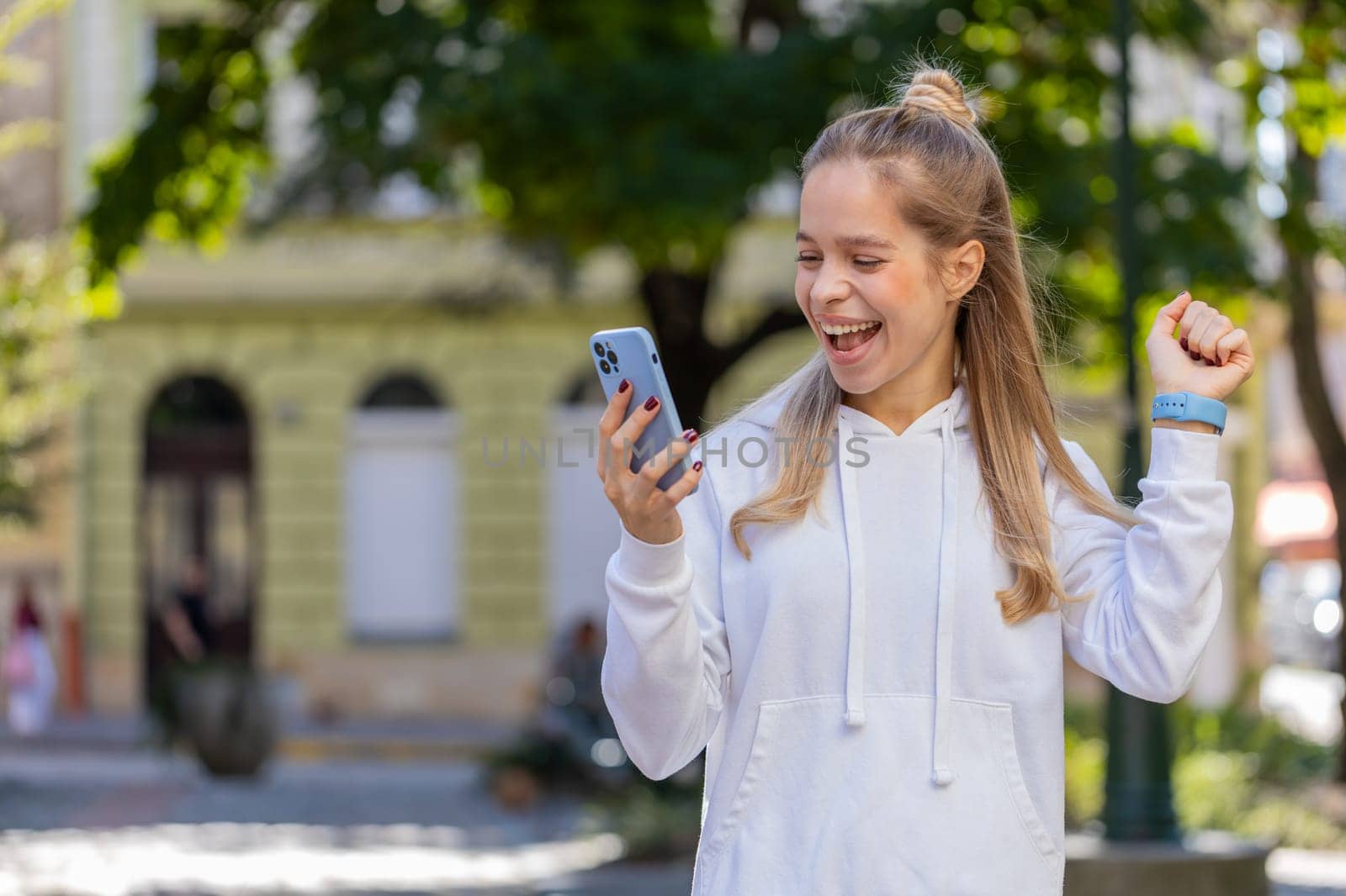 Happy Caucasian young woman using smartphone celebrating win good message news, lottery casino app jackpot victory, online outdoors. Attractive female tourist teenager clenching fists on city street.