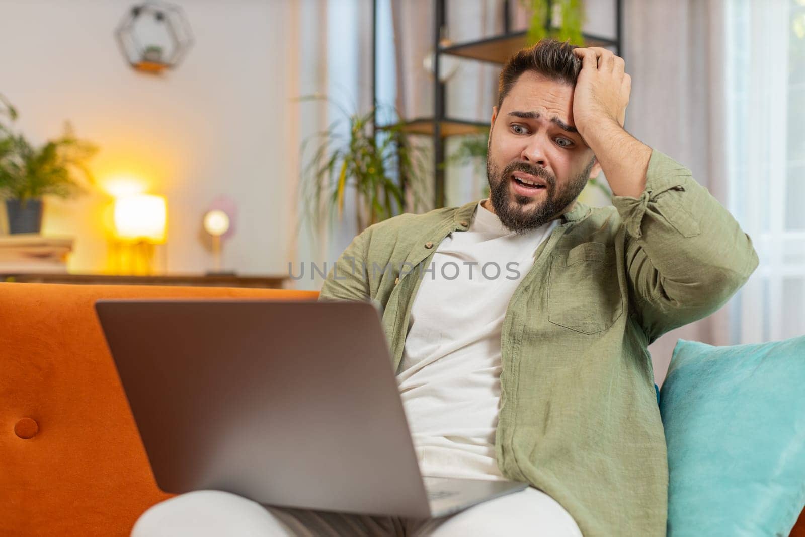 Upset young Caucasian young man sitting on sofa at home using laptop having anxiety and stress. Shocked brunette guy reacting to loss, bad news on netbook in living room apartment. Computer virus.