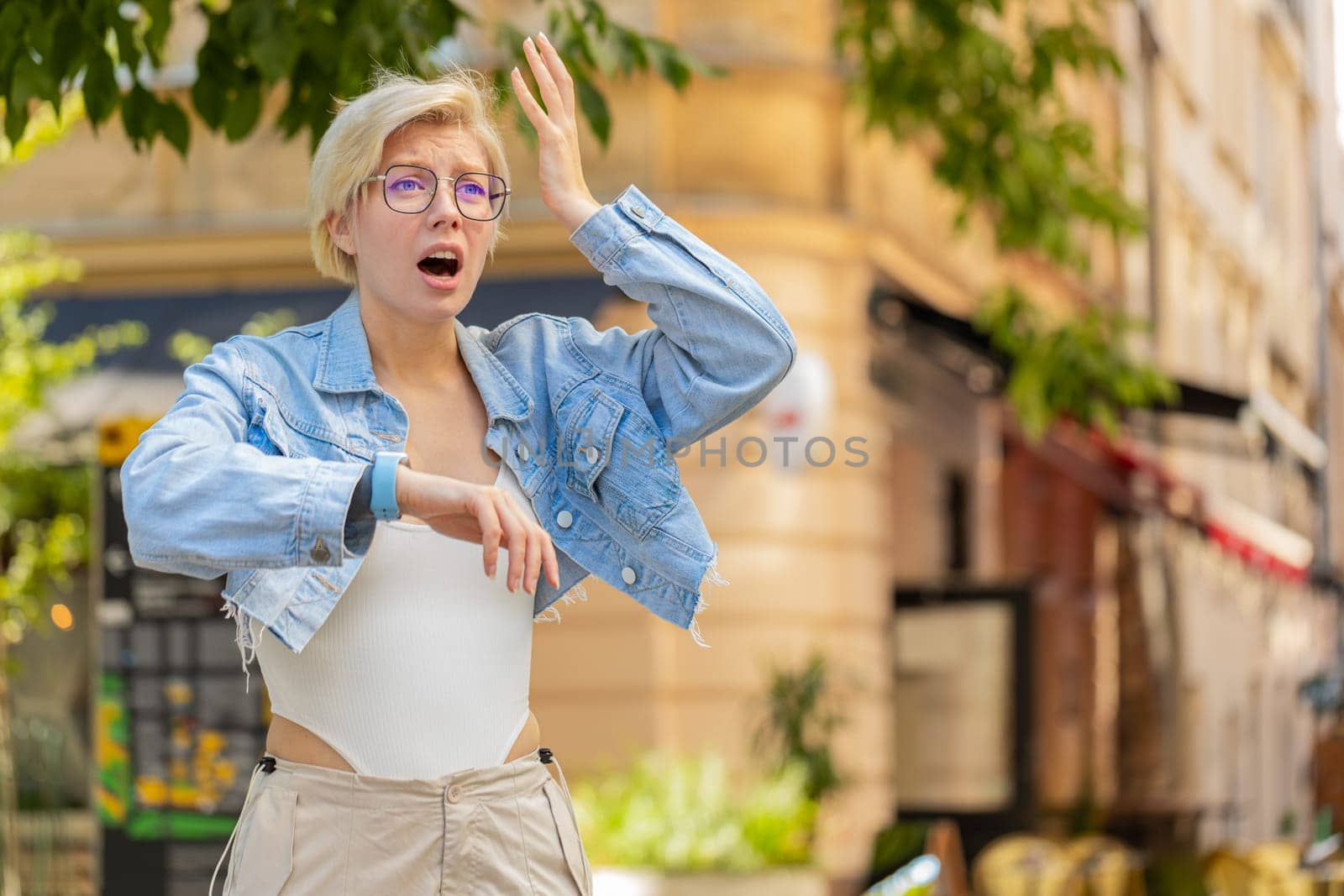 Scared woman worrying to be punctual, with anxiety checking time on watch, running late to work or transport, being in delay, deadline. Blonde girl tourist walking in city street. Town lifestyles.