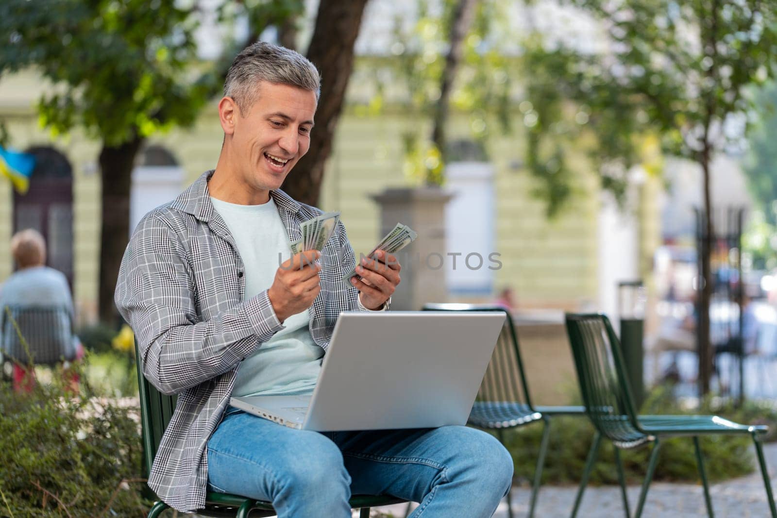 Happy rich man using laptop winner victory counting holding fan of cash money in dollar banknotes sitting on chair city street park. Young male guy tourist planning budget online shopping. Lifestyles