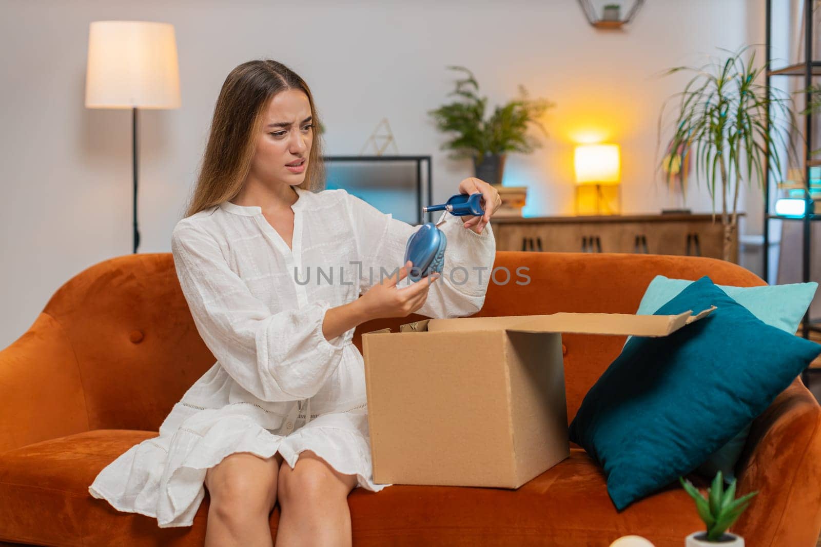 Dissatisfied annoyed female customer opening cardboard box receive damaged broken wireless headphones shocked young woman having problem complaint frustrated with bad order delivery at home. Lifestyle