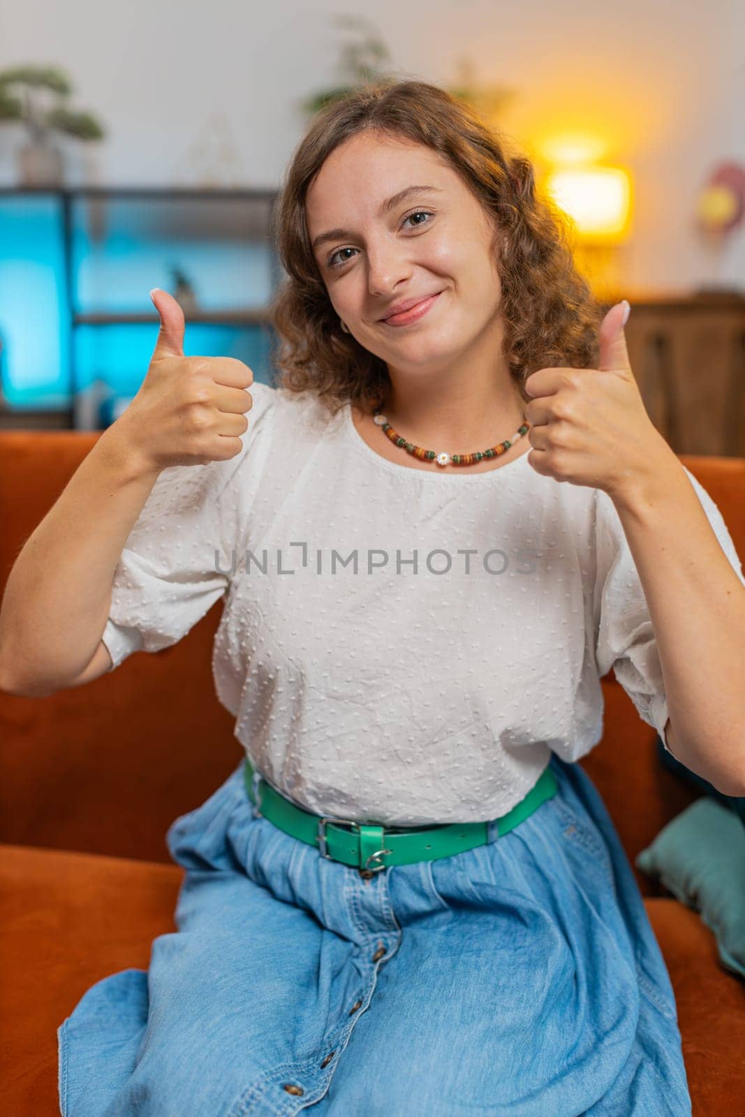 Portrait of excited smiling Caucasian woman looking approvingly at camera showing double thumbs up, like sign, good news, positive feedback. Happy girl sitting on sofa in living room at home. Vertical