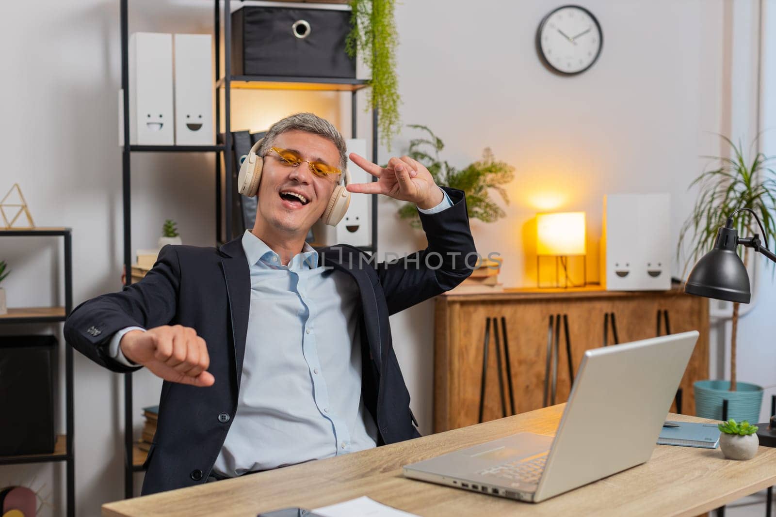 Happy relaxed young businessman working on laptop at office wearing headphones and sunglasses listening favorite energetic disco music, dancing. Freelancer man relaxing, taking a break, celebrating.