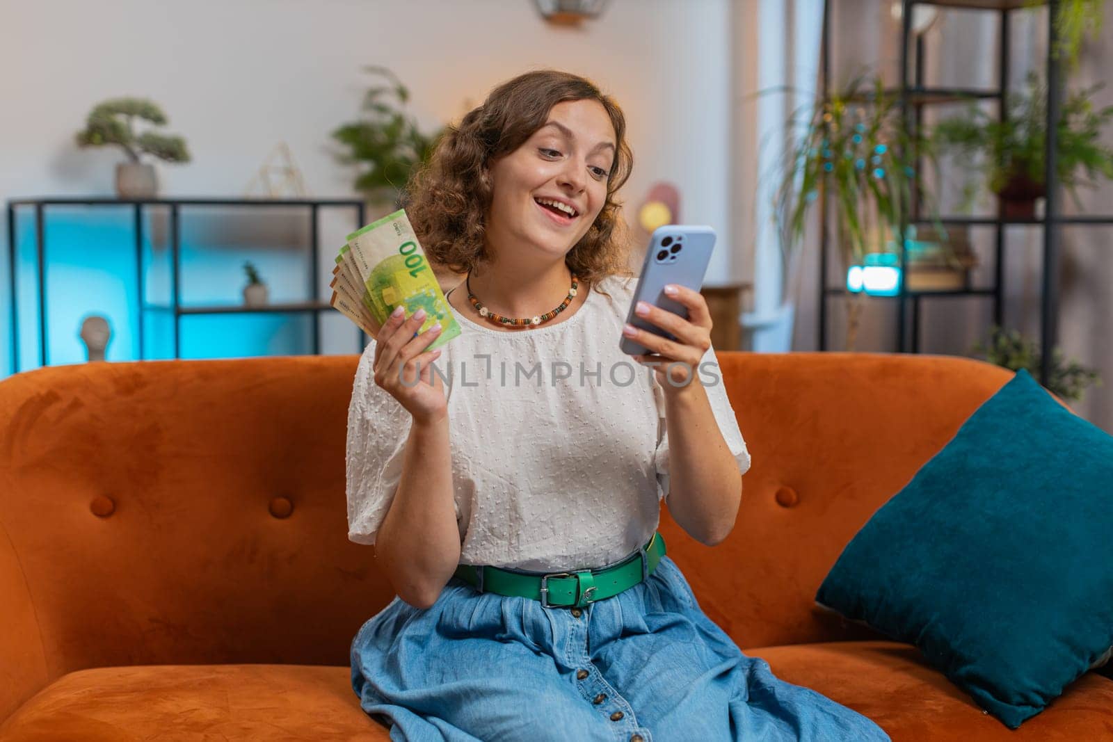 Successful rich woman holding smartphone and waving euro bill money fan sitting on sofa couch at home. Happy girl winning online lottery game planning vacation calculating budget concept. Lifestyles