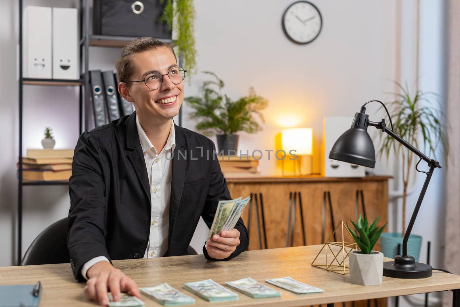 Caucasian successful businessman in formal suit sitting at workplace in home office counting money dollars salary holding stack of dollars, analyzes budget finances. Young freelancer sitting at table.