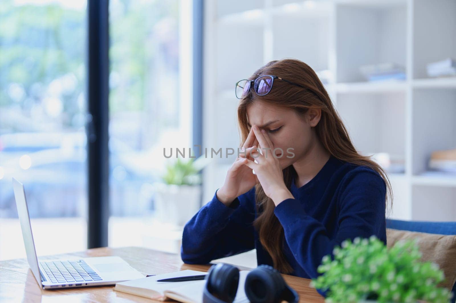 Stressed young woman overwhelmed by e-learning studies at desk with laptop, notebook, headphones, hands clasped to her face in frustration.
