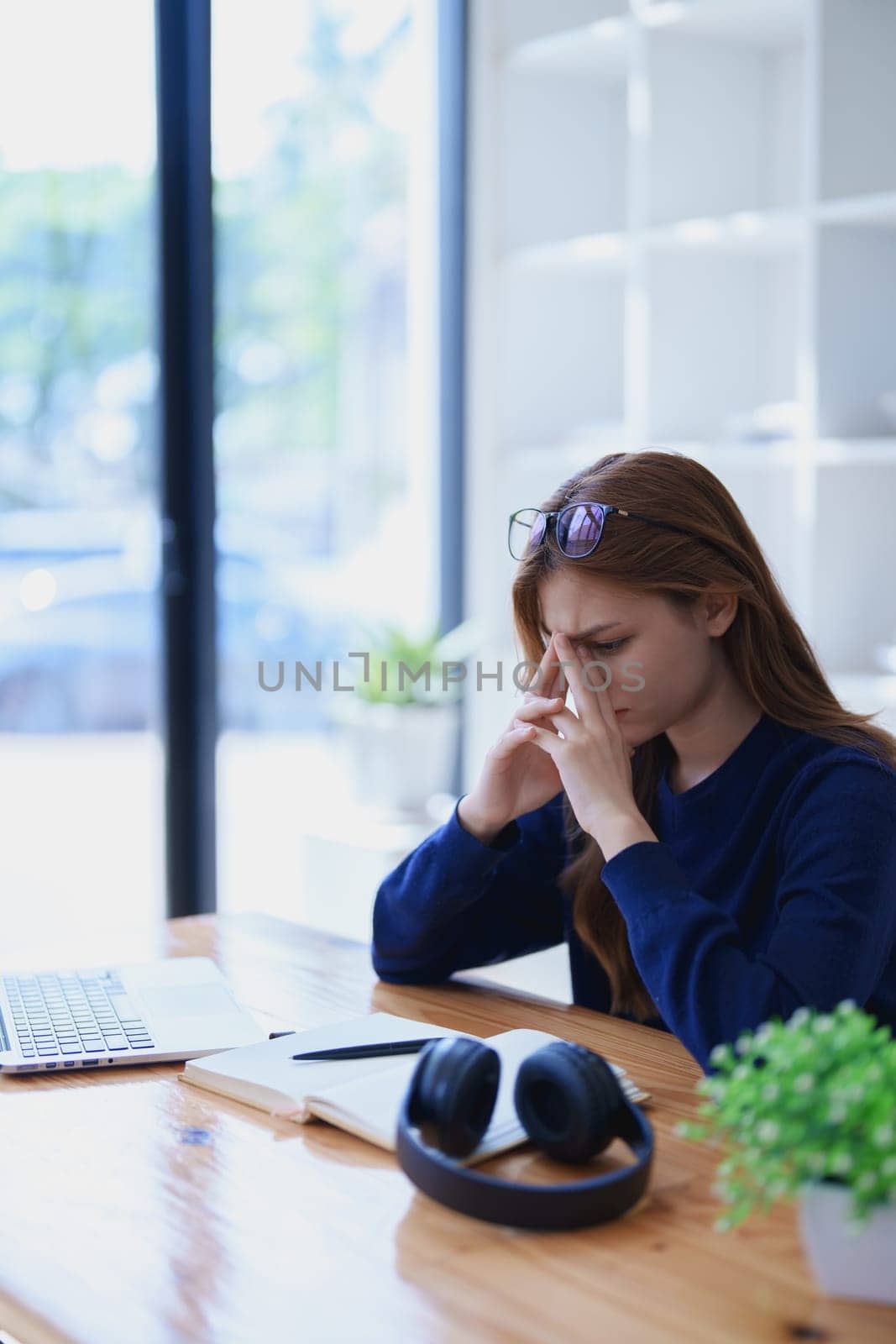 Stressed young woman overwhelmed by e-learning studies at desk with laptop, notebook, headphones, hands clasped to her face in frustration.