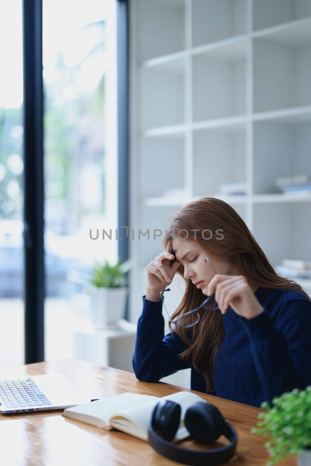 Stressed young woman overwhelmed by e-learning studies at desk with laptop, notebook, headphones, hands clasped to her face in frustration.