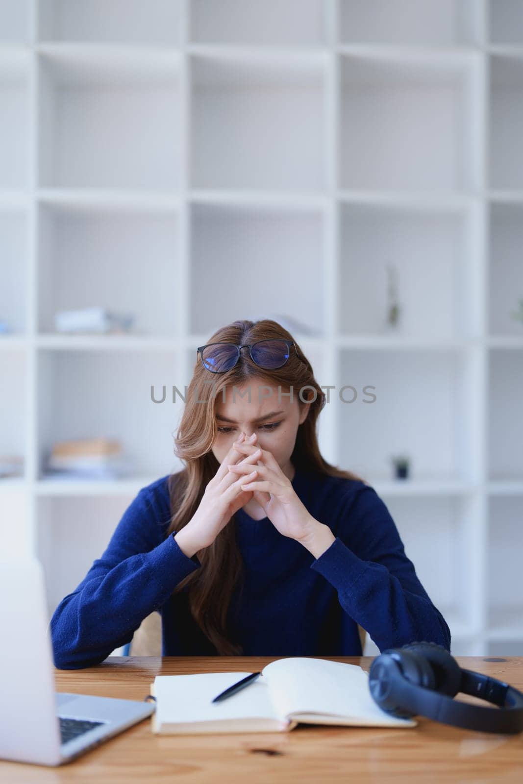 Stressed young woman overwhelmed by e-learning studies at desk with laptop, notebook, headphones, hands clasped to her face in frustration.