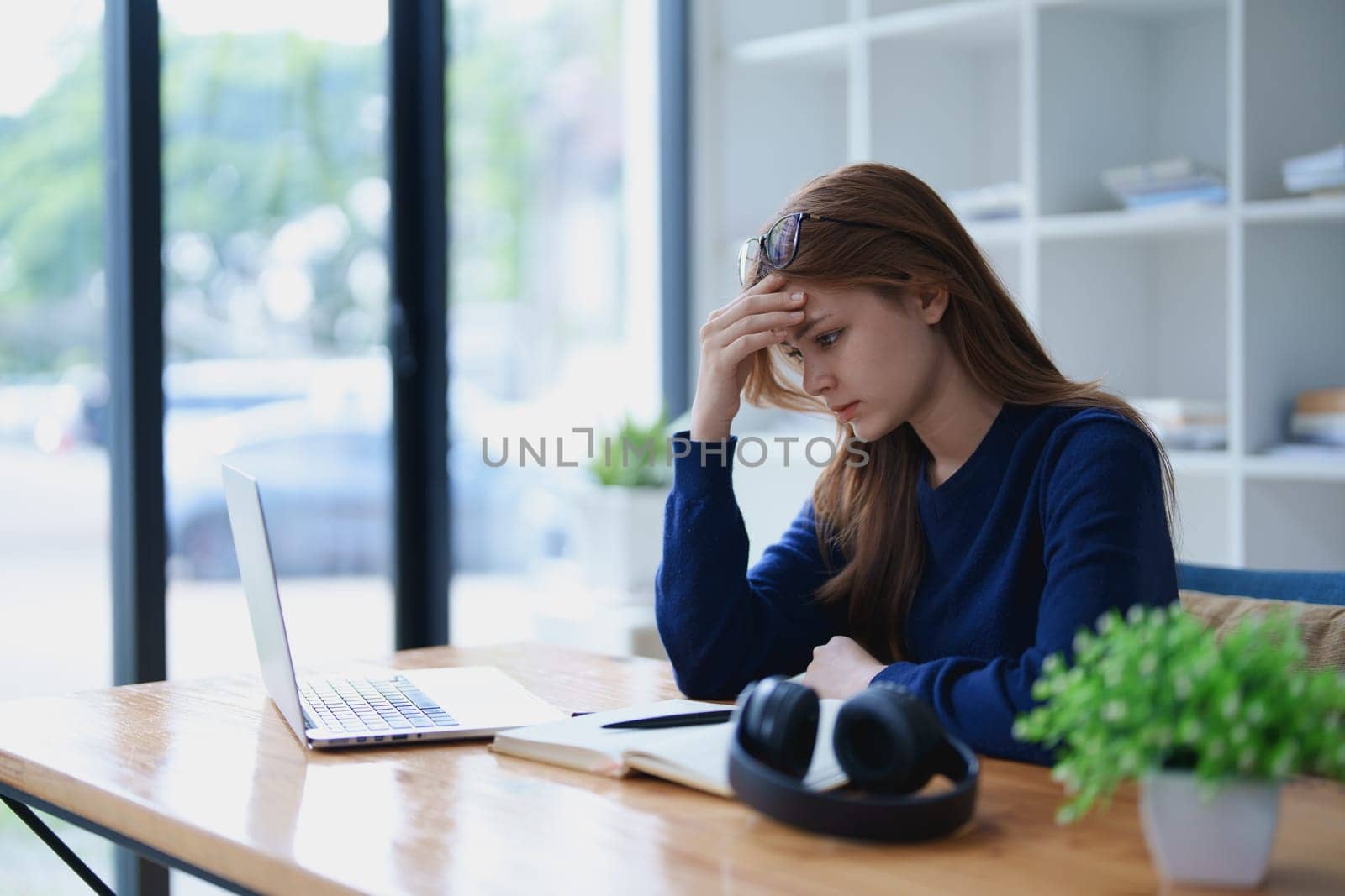 Stressed young woman overwhelmed by e-learning studies at desk with laptop, notebook, headphones, hands clasped to her face in frustration.