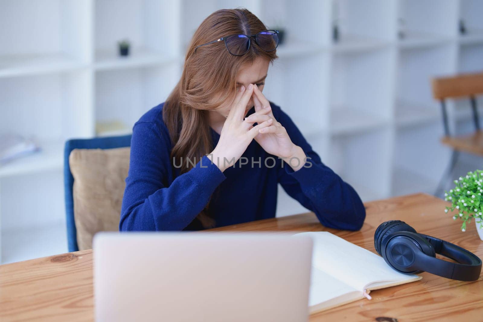 Stressed young woman overwhelmed by e-learning studies at desk with laptop, notebook, headphones, hands clasped to her face in frustration.