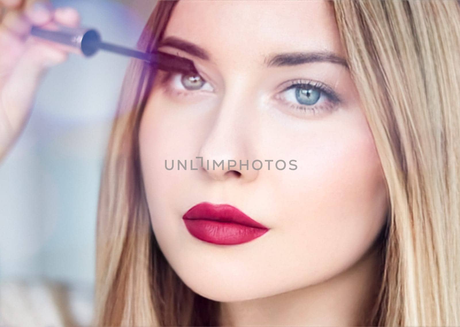 Woman applying mascara with confidence, defined brows and red lipstick. Blonde beauty focused on her eye makeup, cosmetics look