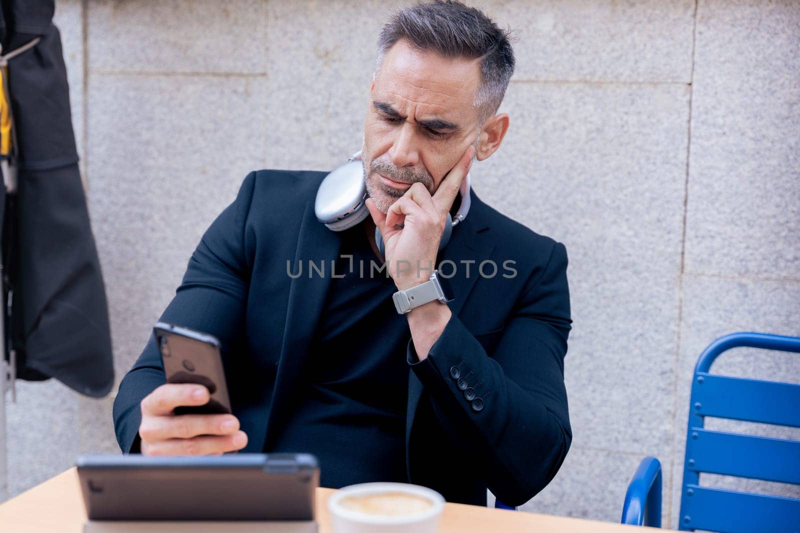 Caucasian mature man with headphones around neck looks thoughtful while checking smartphone. Seated at outdoor cafe with tablet and coffee, reflecting a modern and focused work lifestyle