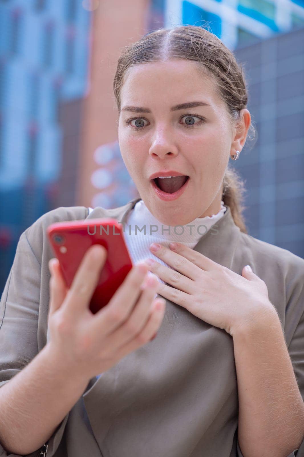 Caucasian woman looking at her smartphone with a shocked and surprised expression. Unexpected news, social media reaction, and technology concept in an urban environment with modern buildings.Vertical