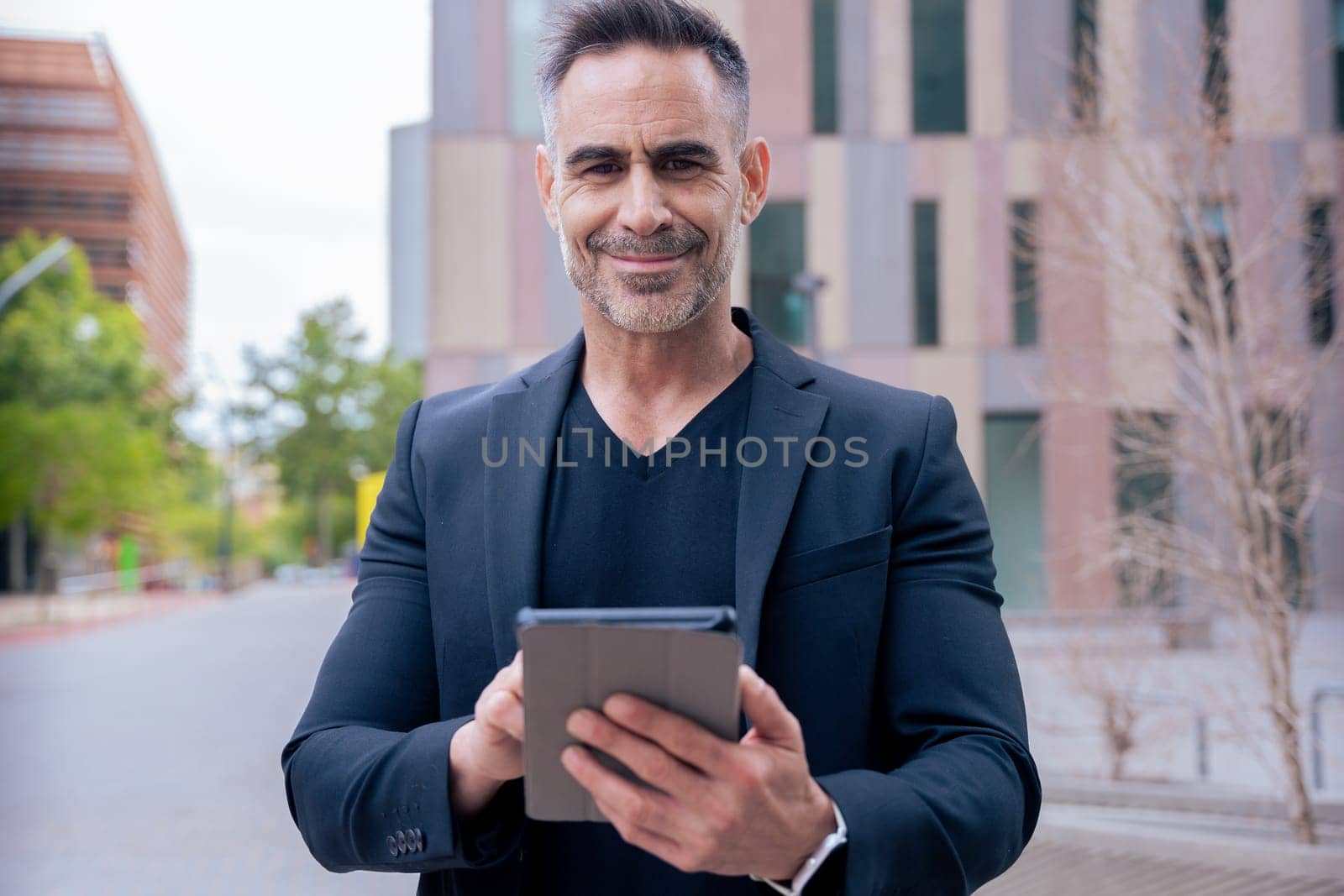 Confident mature businessman working on tablet outside a modern office building. looking at camera. Urban setting Ideal for business, technology, productivity, and professional lifestyle themes