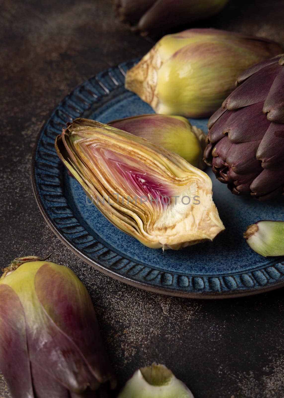 Blue plate full of purple raw romanesco artichokes on dark rustic table closeup. Fresh vegetables bought on farmers market ready to cook. Italian cousine ingredient