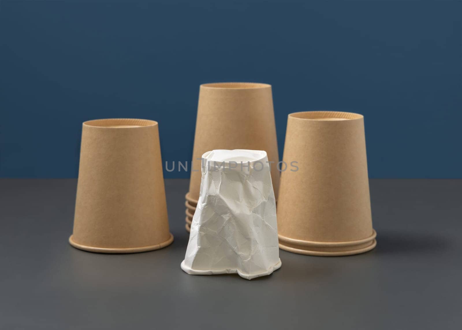 Disposable beige kraft paper cups and crumpled used white one on gray against blue closeup, mockup, copy space. Empty cardboard coffee cups. Consumerism concept