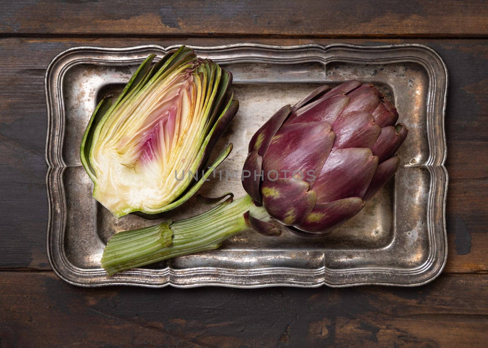 Half and whole purple raw roman artichokes on old metal tray on dark weathered wooden table top view. Fresh vegetables bought on farmers market ready to cook. Italian cousine ingredient