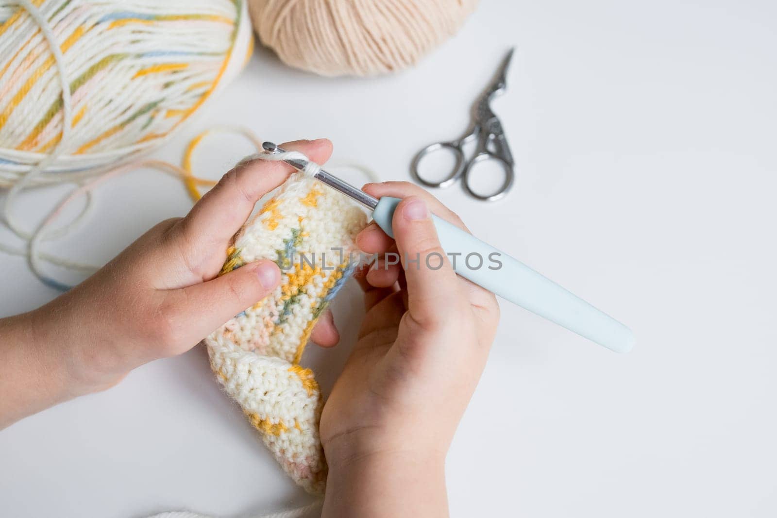 Child crocheting colorful yarn with hook and scissors on table.