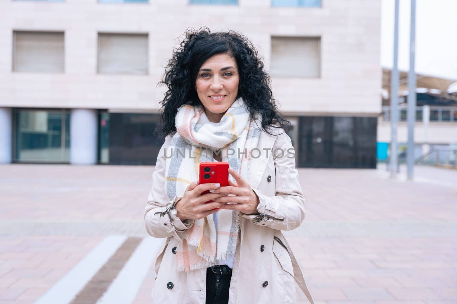 Happy caucasian woman standing outdoors in urban setting, wearing coat and scarf, using her red smartphone. She is smiling and relaxed looking at camera