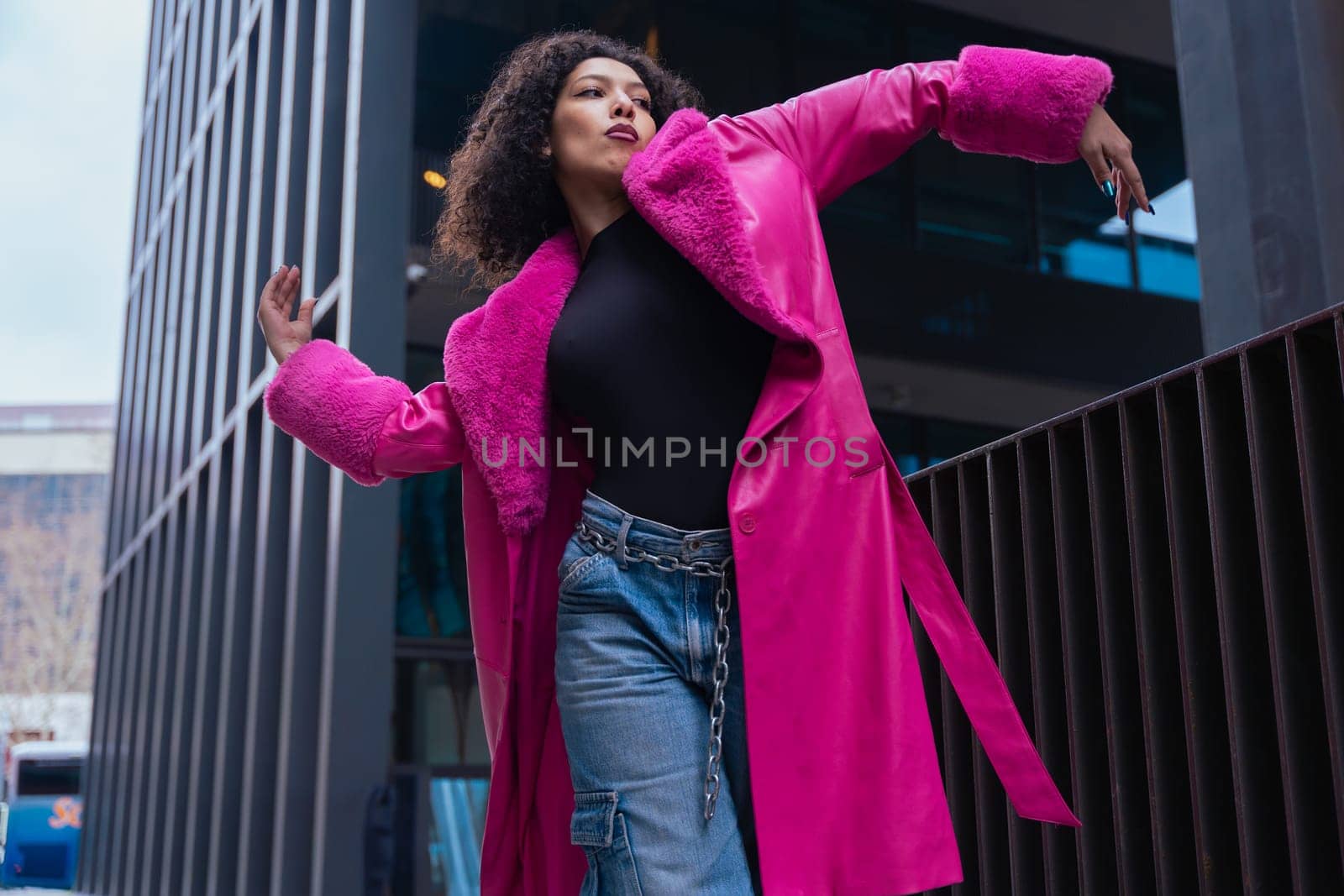 African american dancer with curly hair wearing a pink coat, expressing movement and energy in an urban environment. Modern fashion, contemporary dance,and artistic performance against a city backdrop