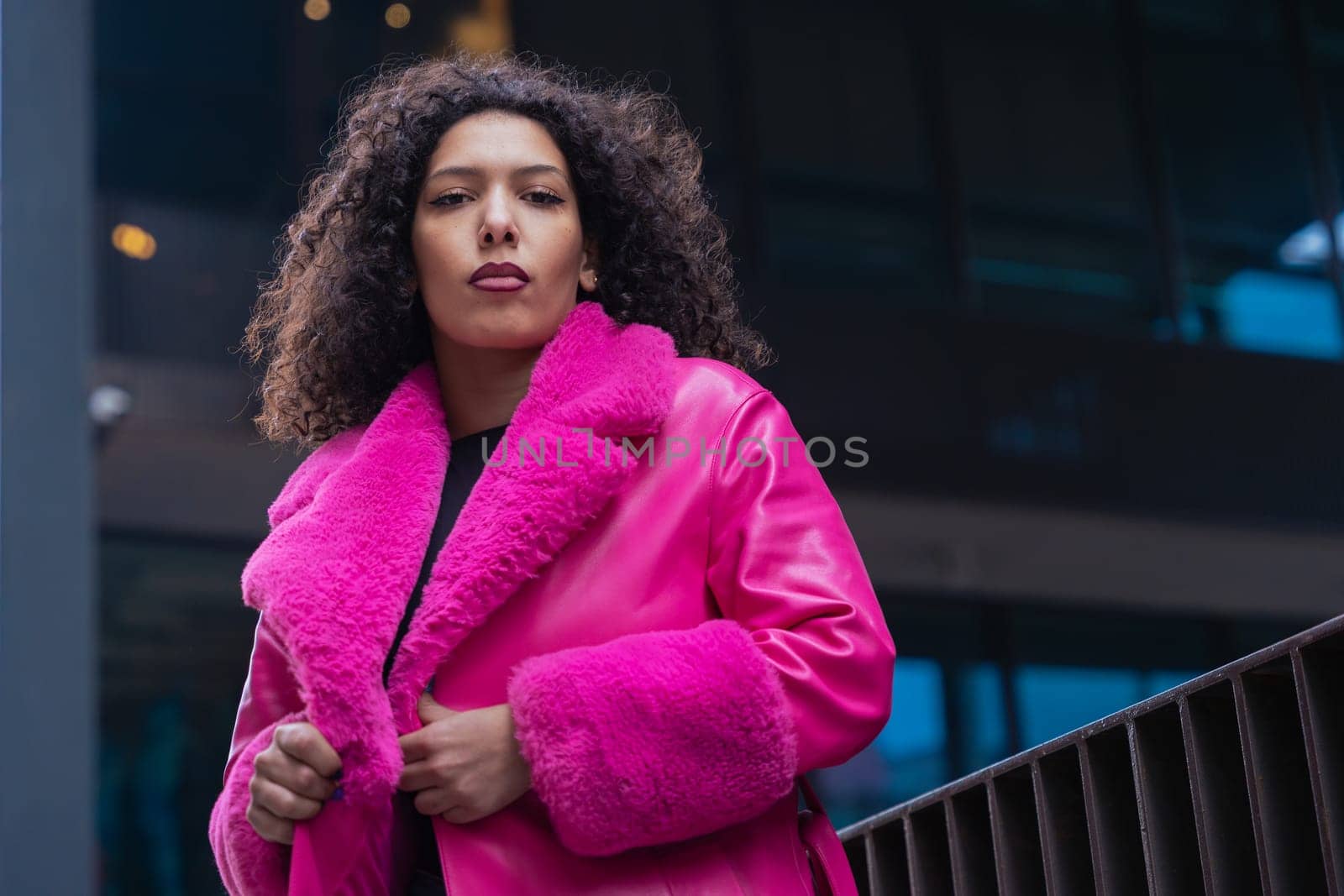 Horizontal portrait of a fashionable young woman in a bold pink coat and jeans, walking confidently through a modern urban area. Her serious expression adds looking at camera