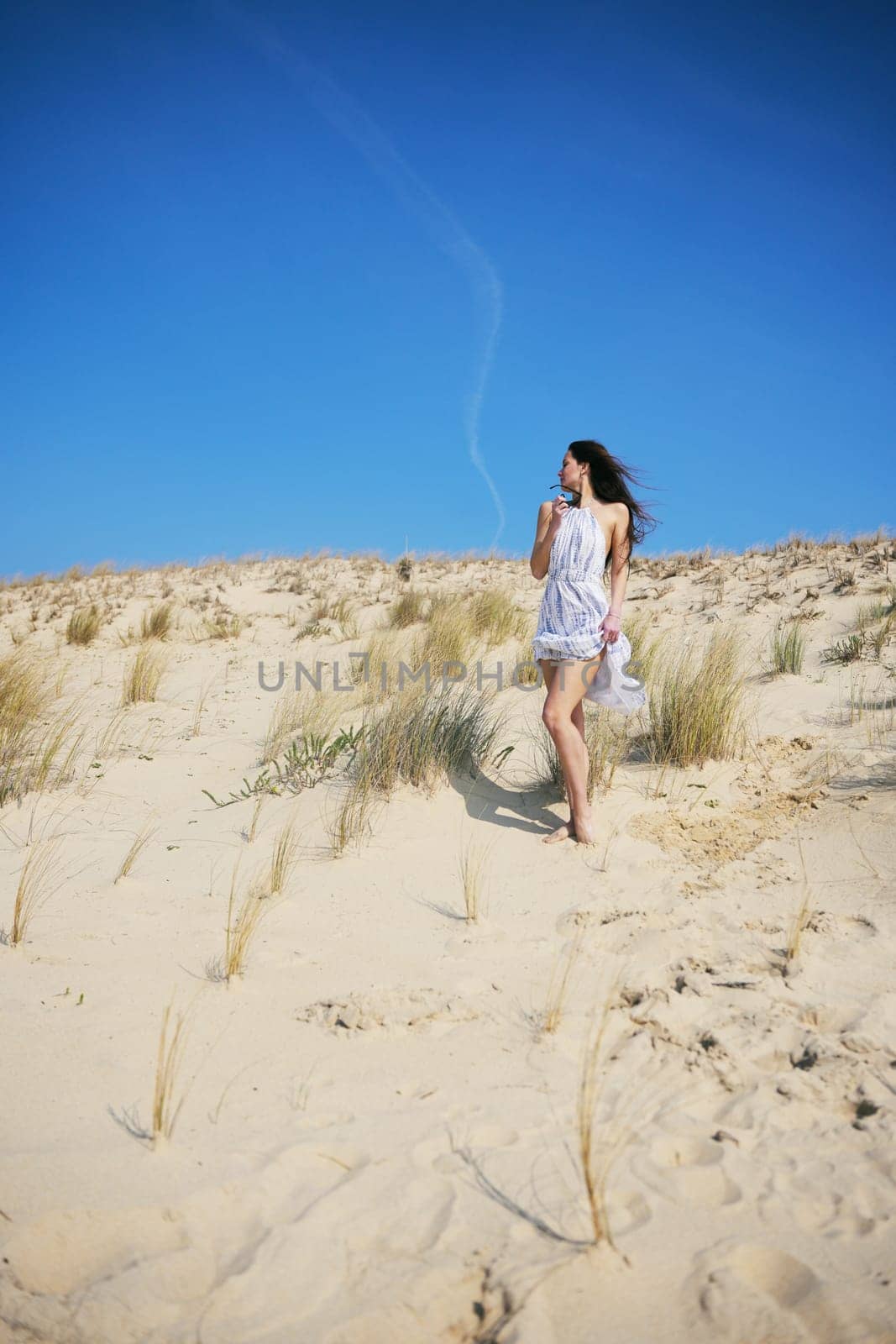 A young woman in a flowing white summer dress walks barefoot across a sunlit sand dune, surrounded by dry grass under a vivid blue sky, capturing a sense of freedom, peace, and wanderlust.