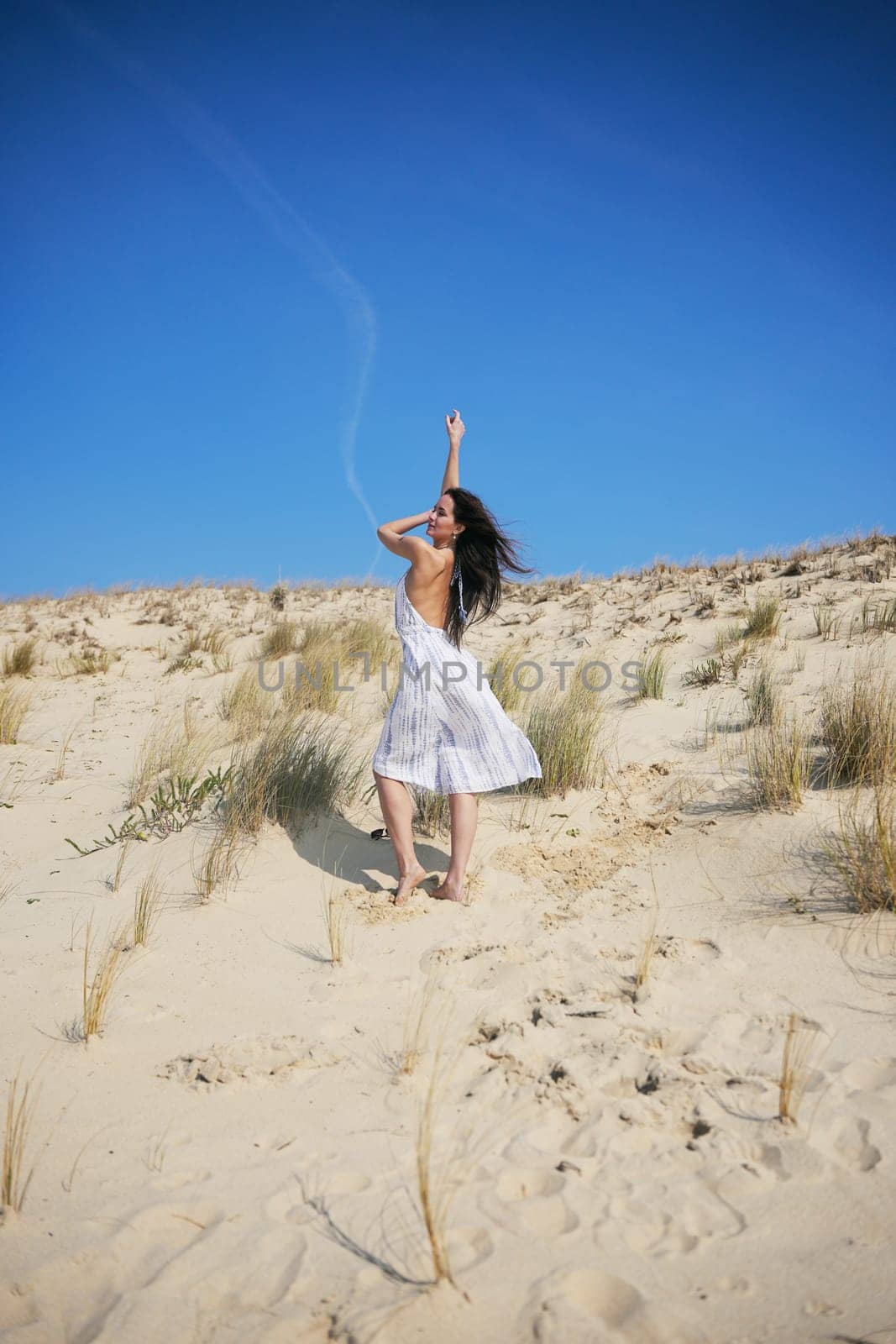 A stylish woman poses on a golden dune in a breezy summer dress. Her carefree posture and windblown hair highlight effortless beach fashion.