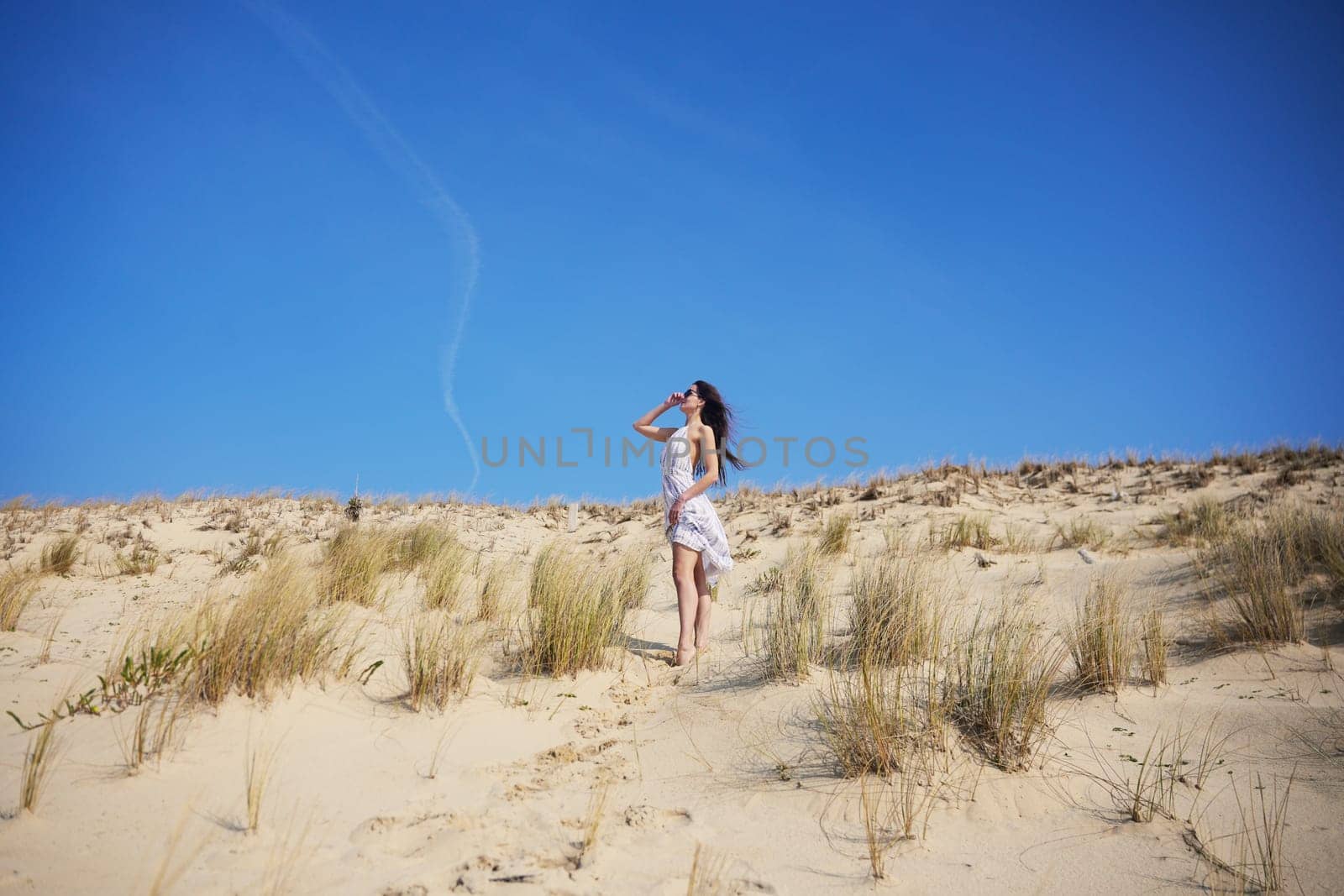 A young woman in a flowy white dress walks alone across a peaceful sand dune at sunrise, surrounded by soft light, dry grass, and open skies evoking serenity, freedom, and calm adventure.