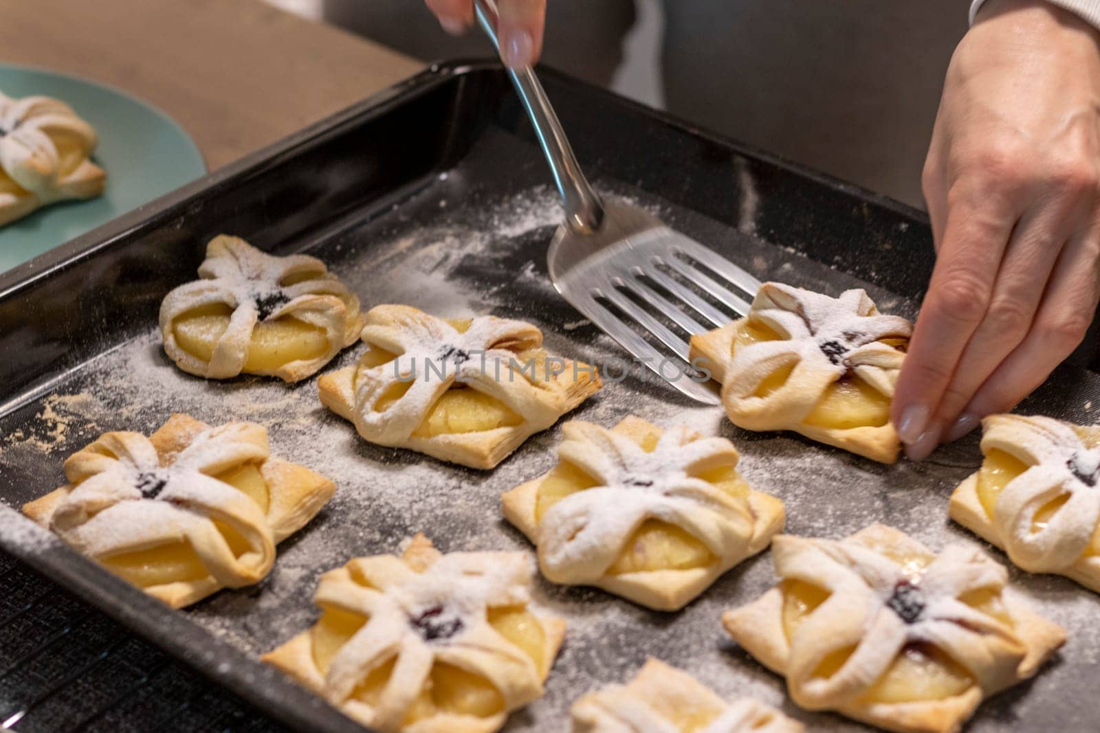 A woman pick the fruit puff pastries with spatula from baking tray and places them on ceramic plate
