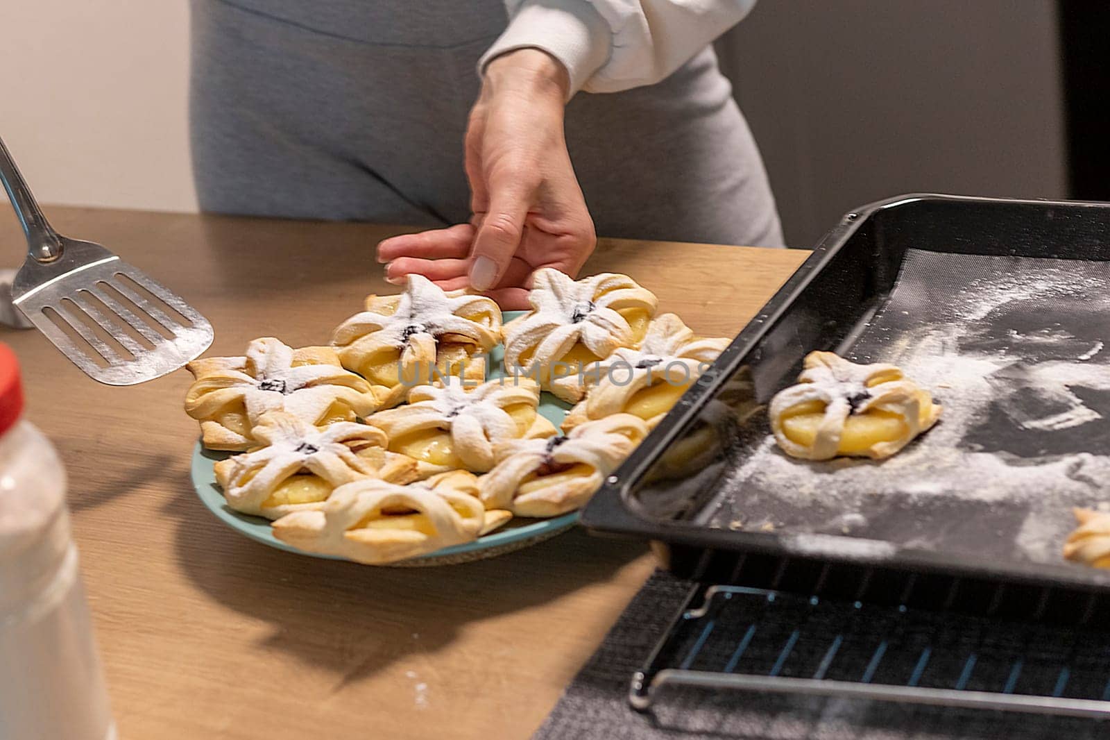 A woman pick the fruit puff pastries with spatula from baking tray and places them on ceramic plate.