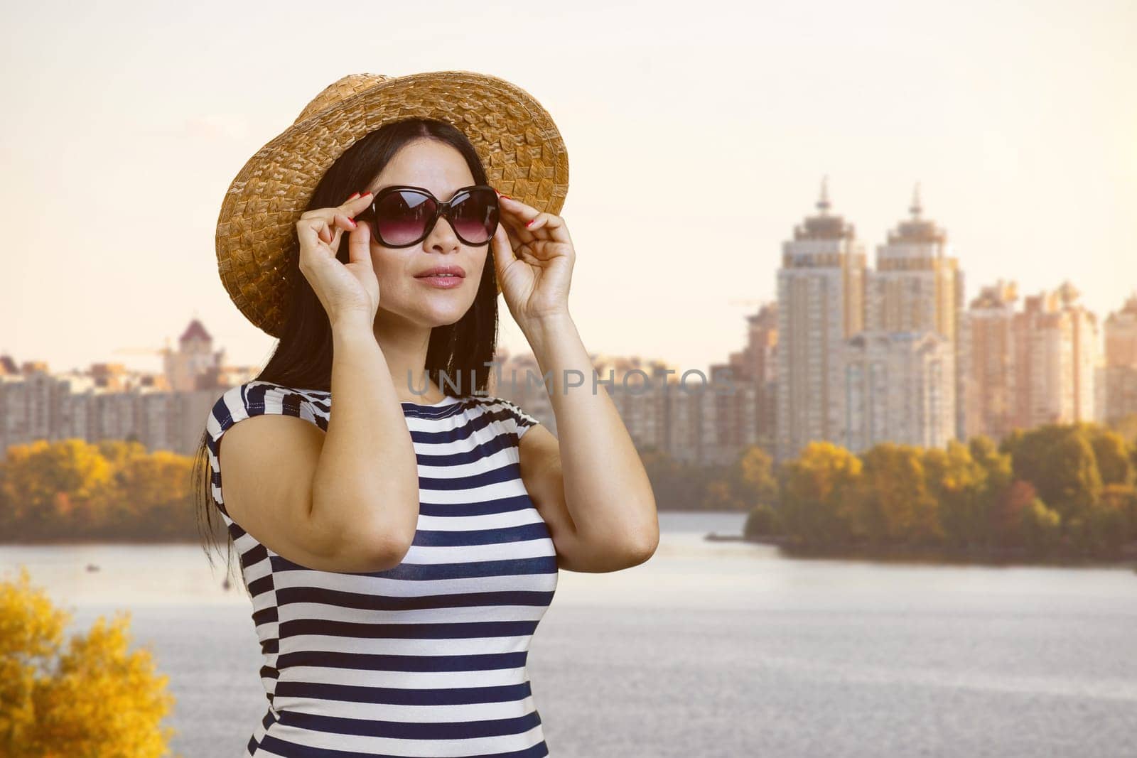 Young brunette woman puts on sunglasses outdoors. City river background.