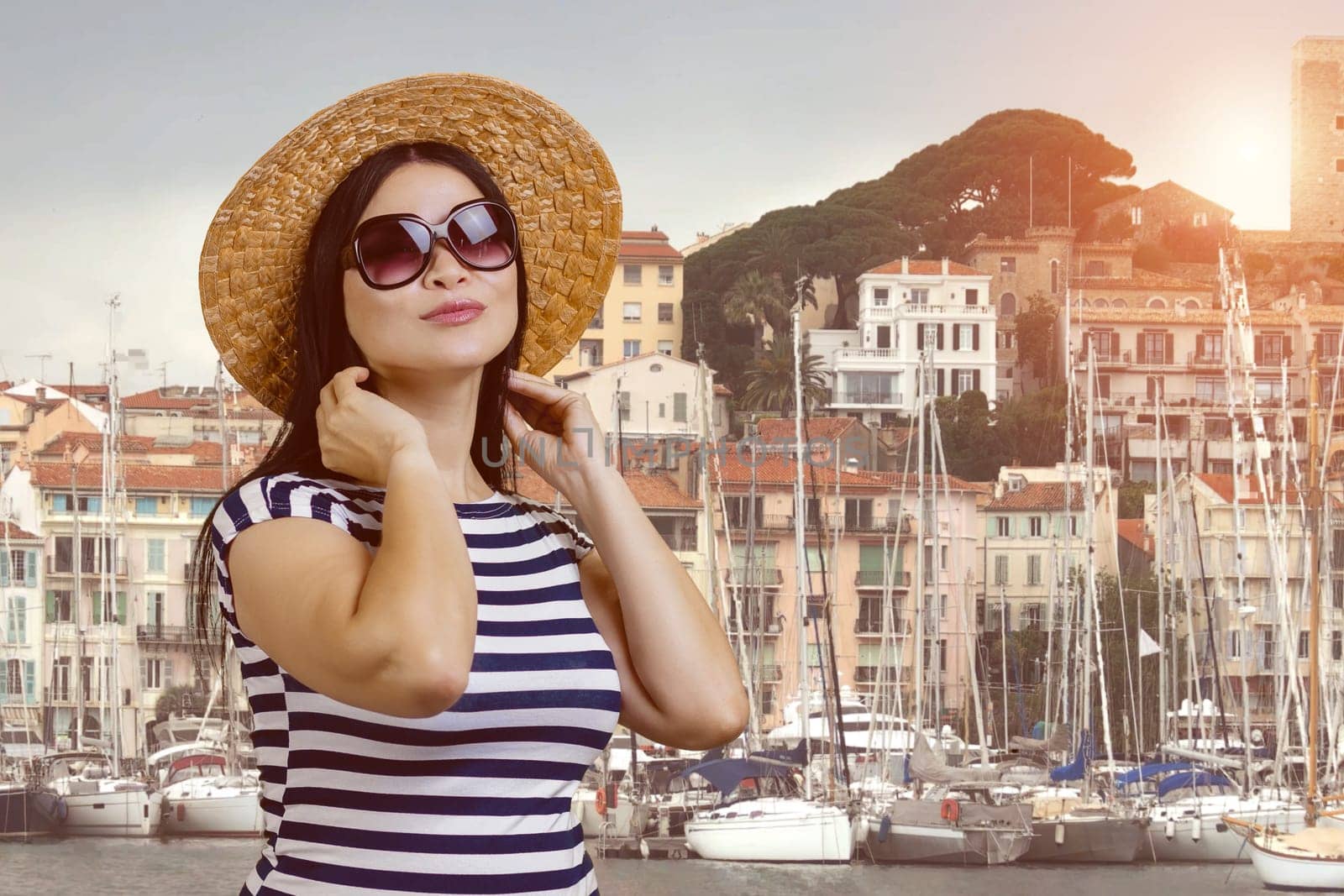 Young brunette female tourist in sunglasses and straw hat is posing outdoors. Yacht harbour and small european houses on the background.