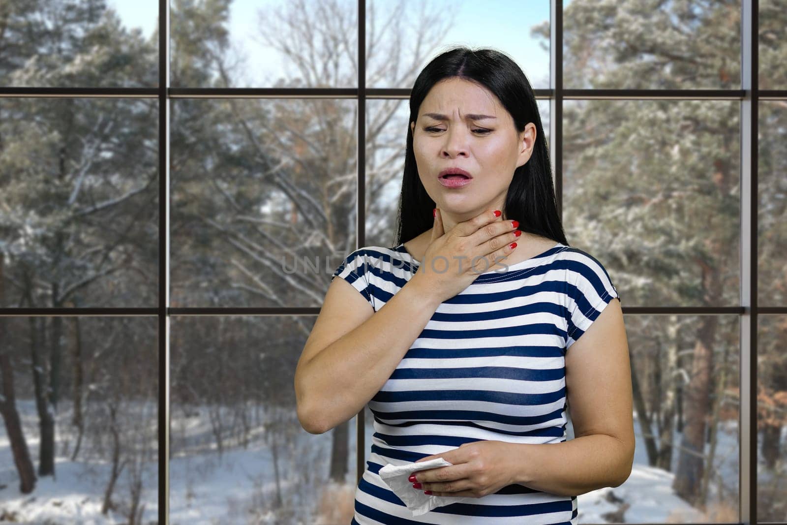 Young asian woman is suffering from cold touching her neck and holding a napkin. Ckeckered windows backgroud with view on a winter landscape.