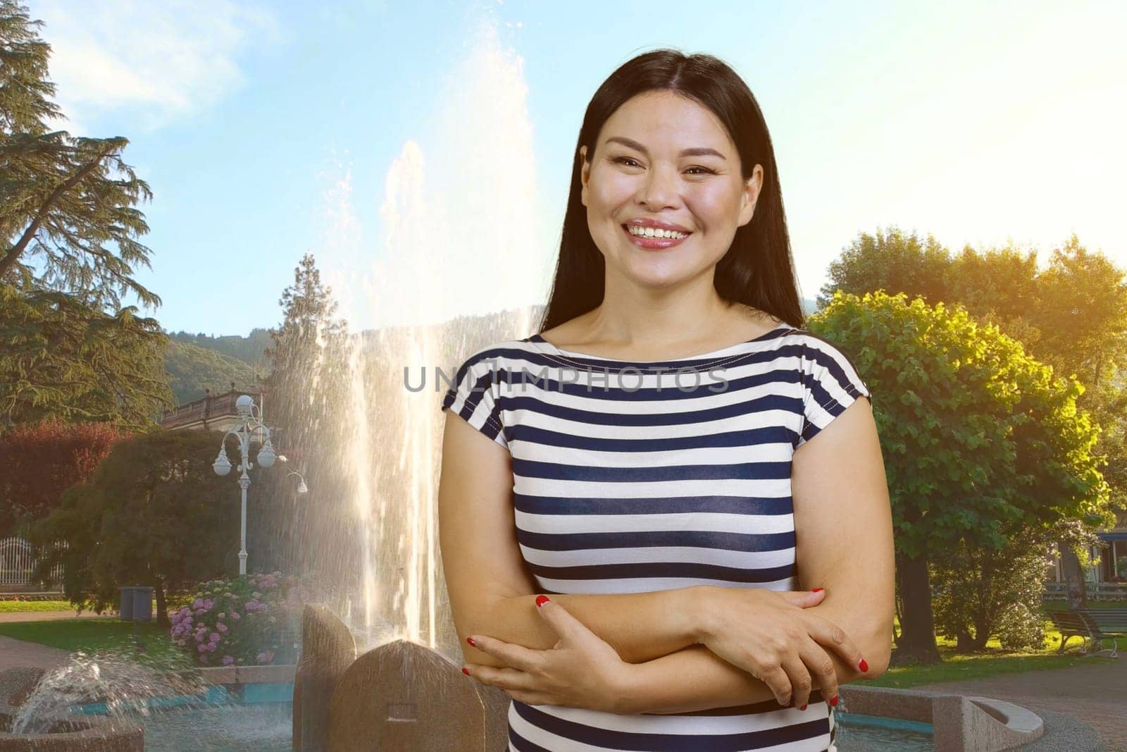 Portrait of cheerful asian woman with crossed hands standing outdoors. Park fountain background.