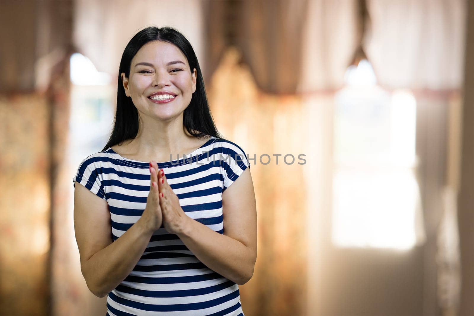 Happy cheerful asian woman with praying hands gesture. Standing indoors on windows backgound.