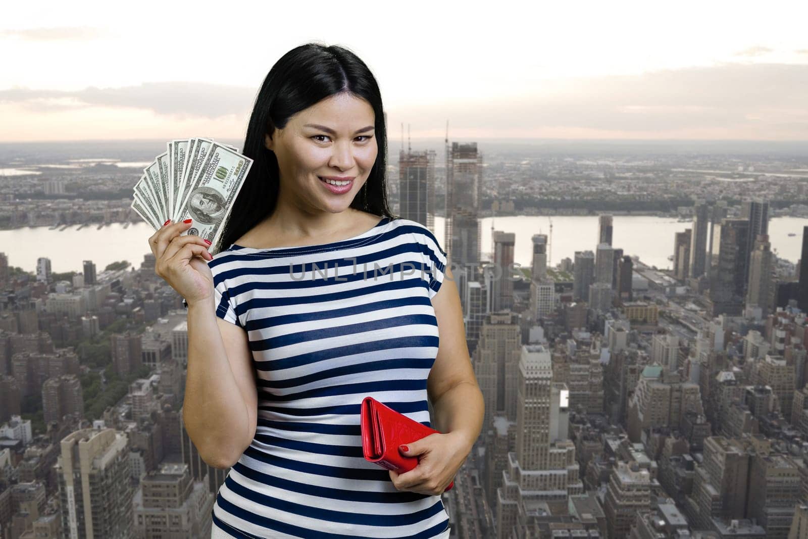 Young asian woman is holding fan of dollars from a red wallet. Urban cityscape background.