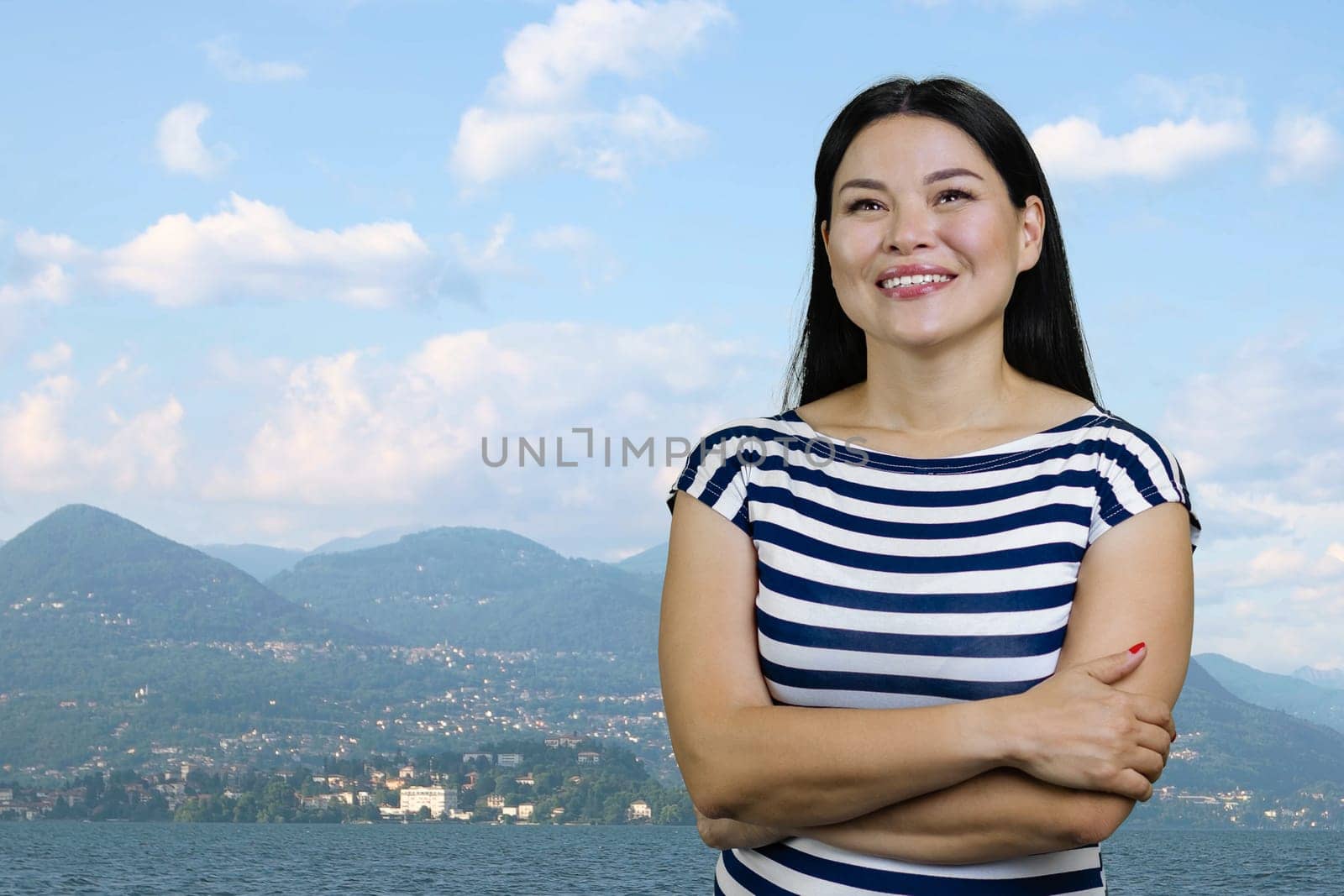 Young asian cheerful pensive woman with folded arms standing outdoors. Mountains and cloudy sky background.