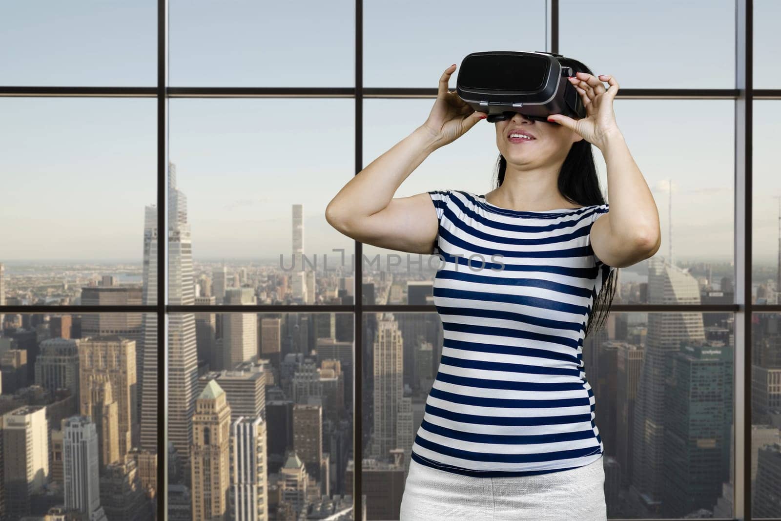 Happy woman getting VR experience in headset glasses. Checkered windows background with cityscape view.