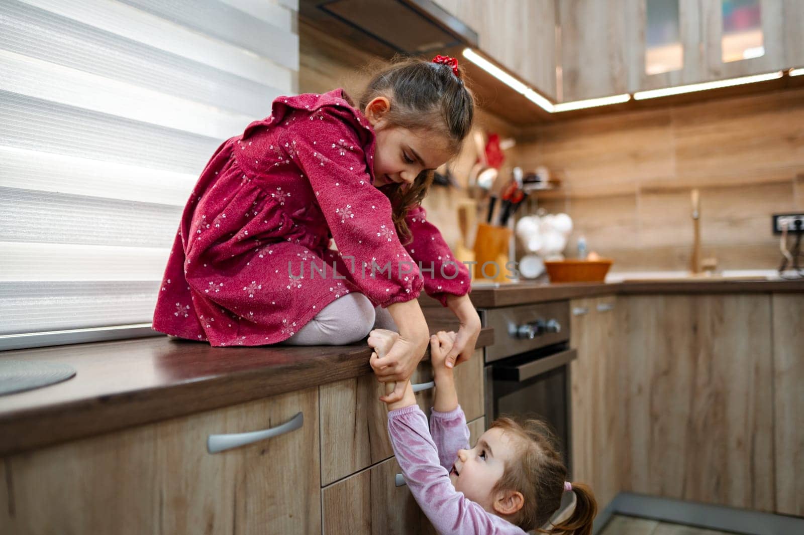 Two young sisters display their bond in a cozy kitchen setting. One girl sits joyfully on the counter, while the other playfully tries to pull her down, creating a tender and lively atmosphere.